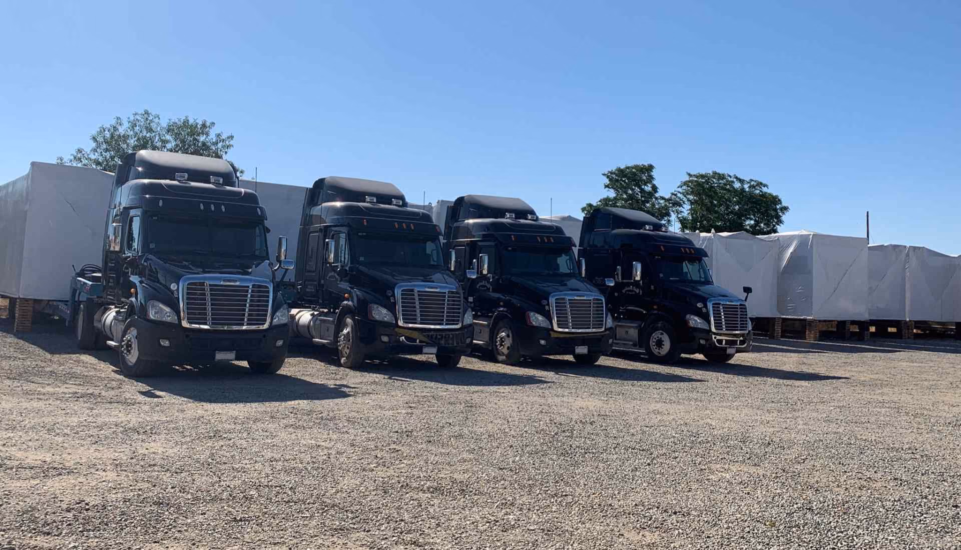 Four black semi trucks parked in a gravel yard beside wrapped modular buildings
