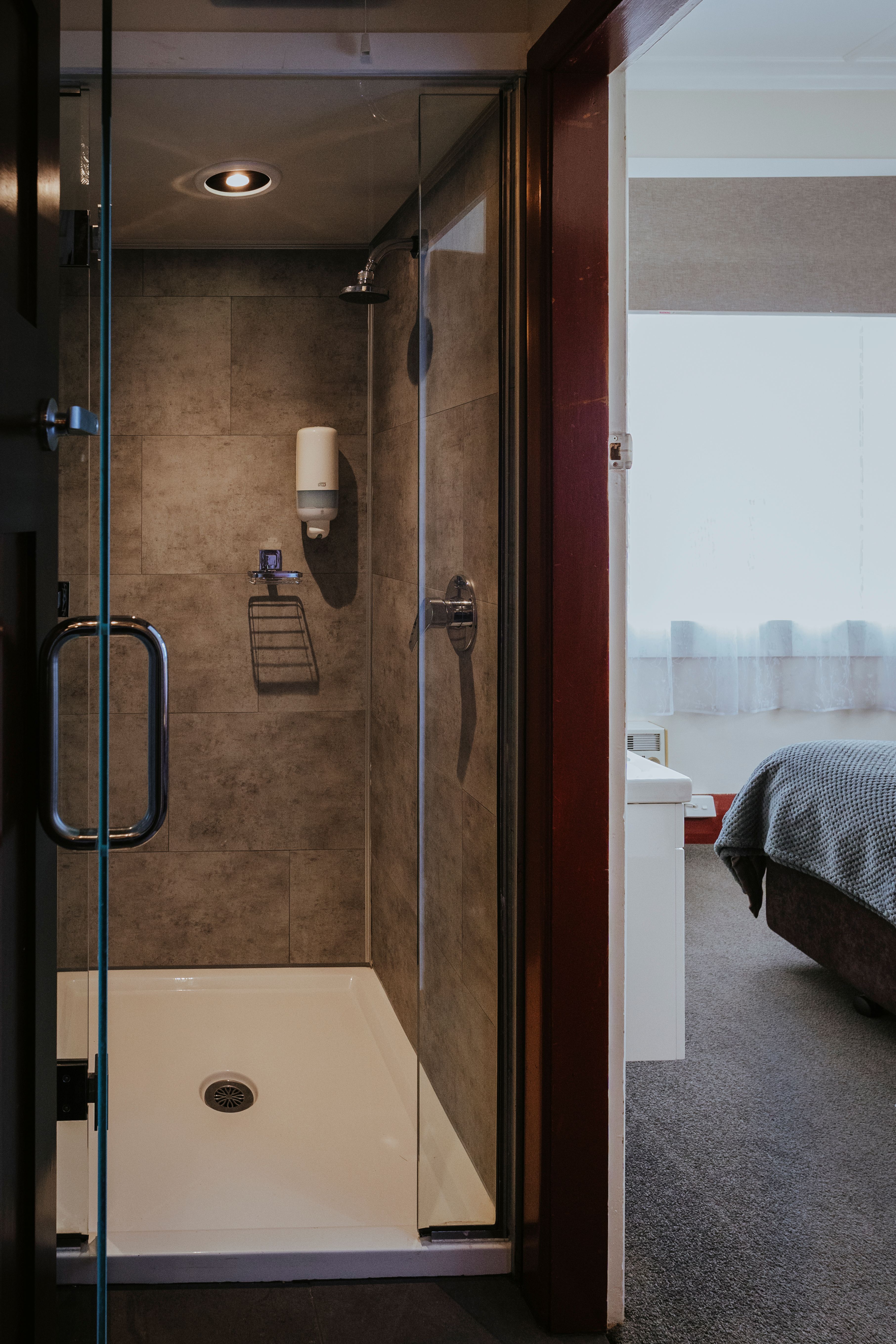 Compact bathroom shower with glass door and stone tile walls next to a bedroom with carpet and a gray blanket on the bed.