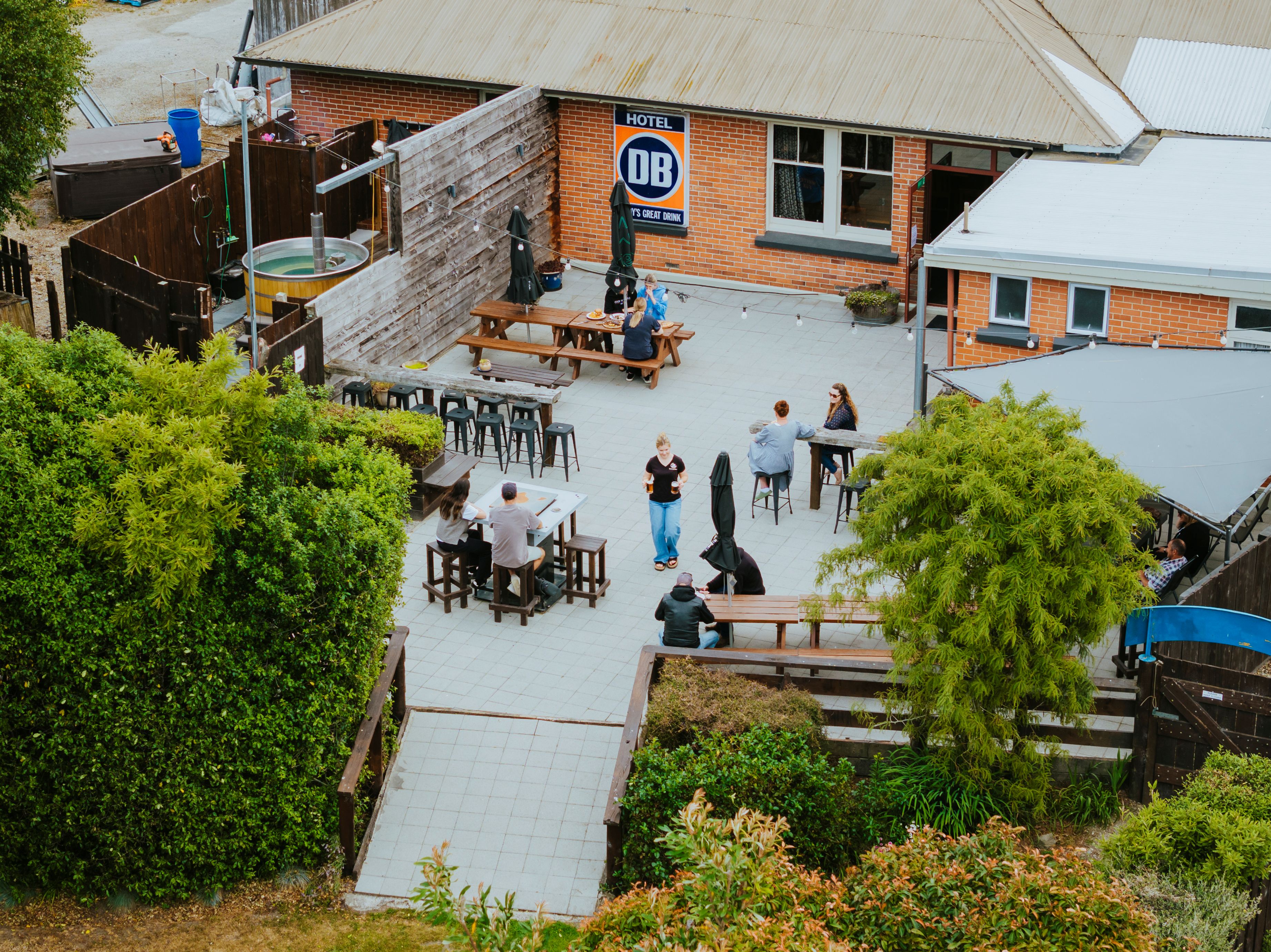 Outdoor patio area with people sitting at wooden tables and benches near a brick building with a DB Hotel sign.