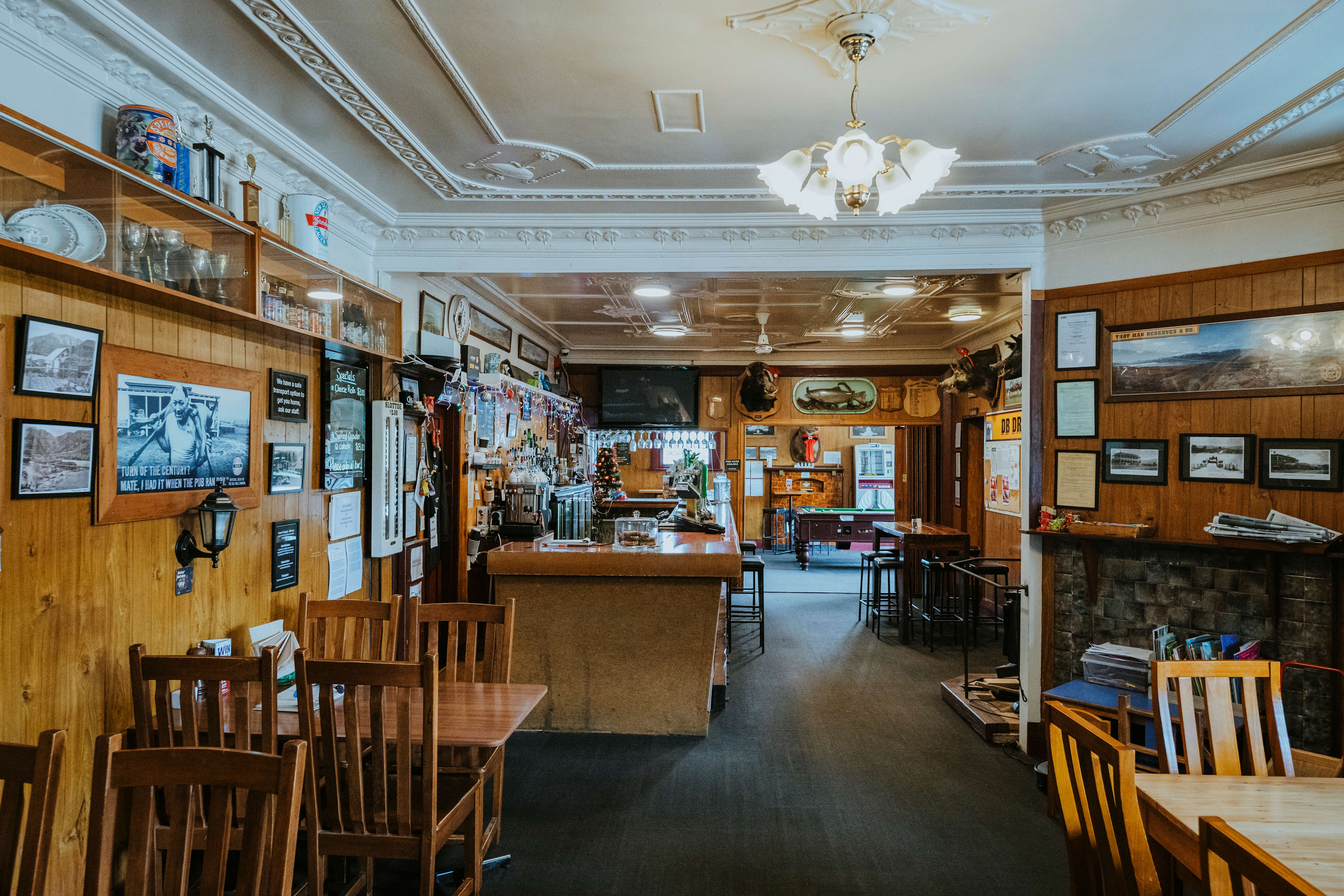 Interior of a cozy vintage pub with wooden tables and chairs, a bar counter, wall-mounted trophies, framed photos, and a pool table in the background.