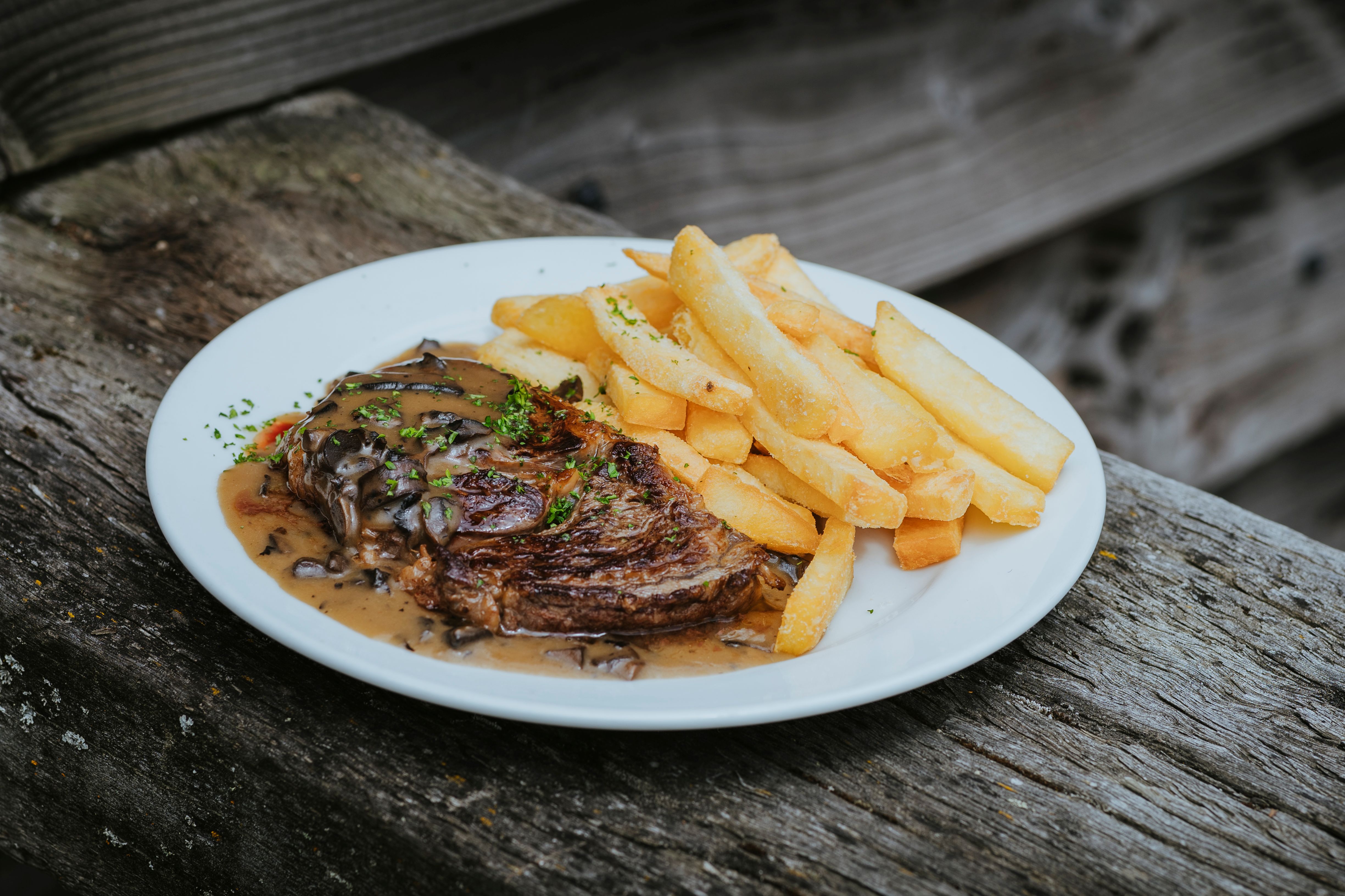 Grilled steak topped with mushroom sauce and parsley served with thick-cut fries on a white plate.