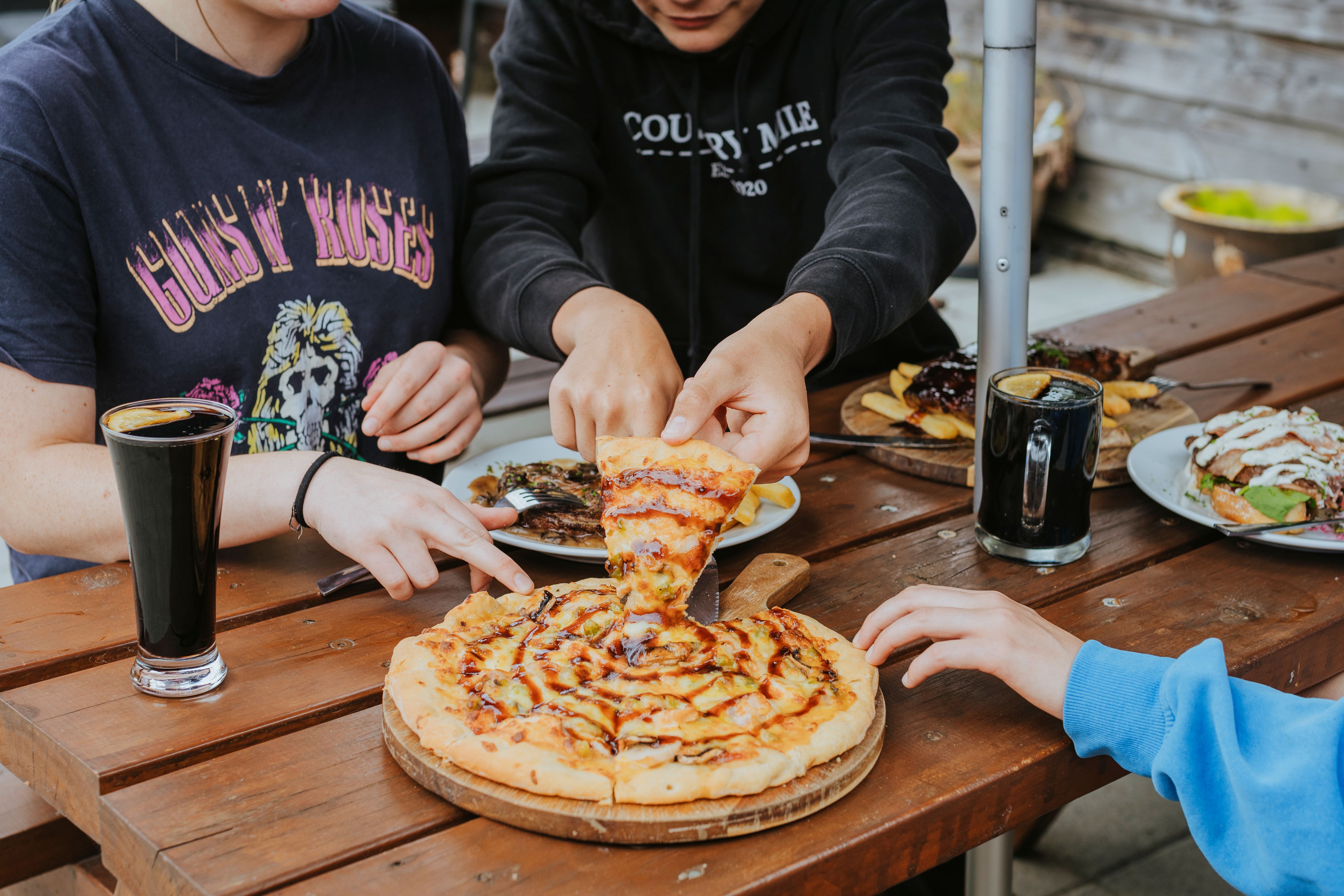 Three people sharing a pizza at a wooden table with drinks and plates of food.