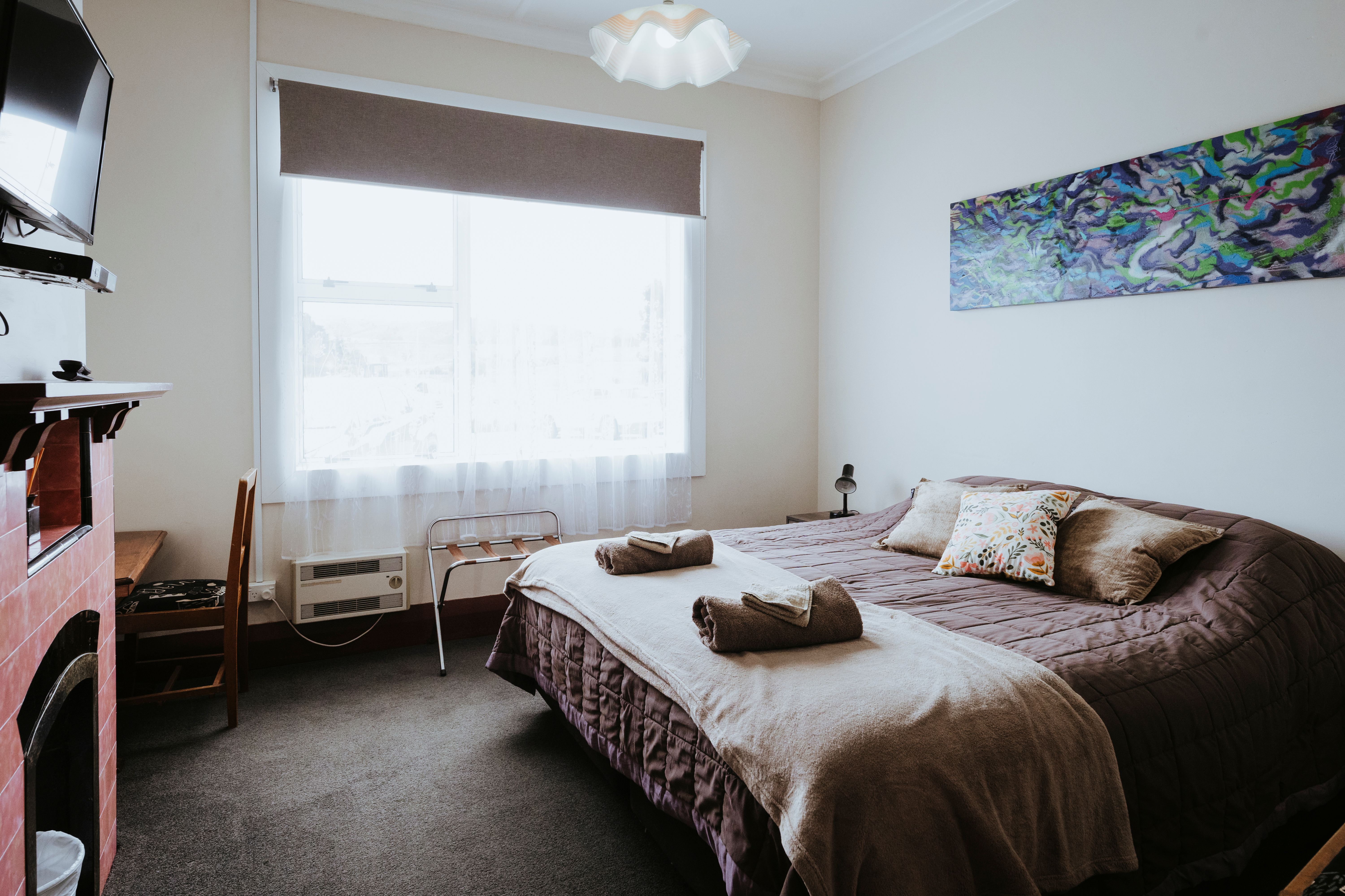 Bedroom with a large bed covered with brown bedding, three pillows, two rolled towels with washcloths, a patterned artwork on the wall, and a window with brown blinds.