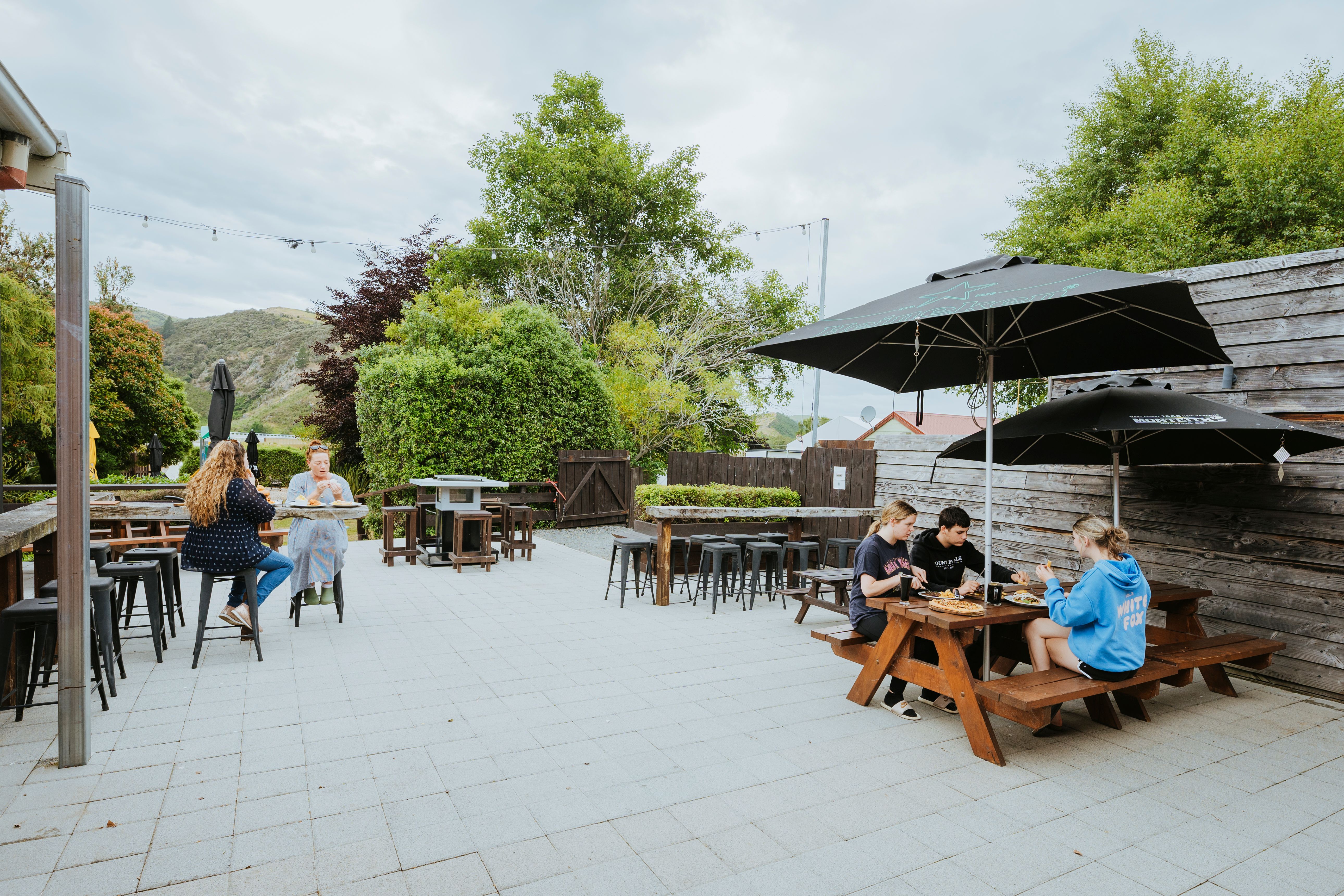 Outdoor beer garden with wooden picnic tables and shaded umbrellas where six people are eating and socializing surrounded by green trees and hills.