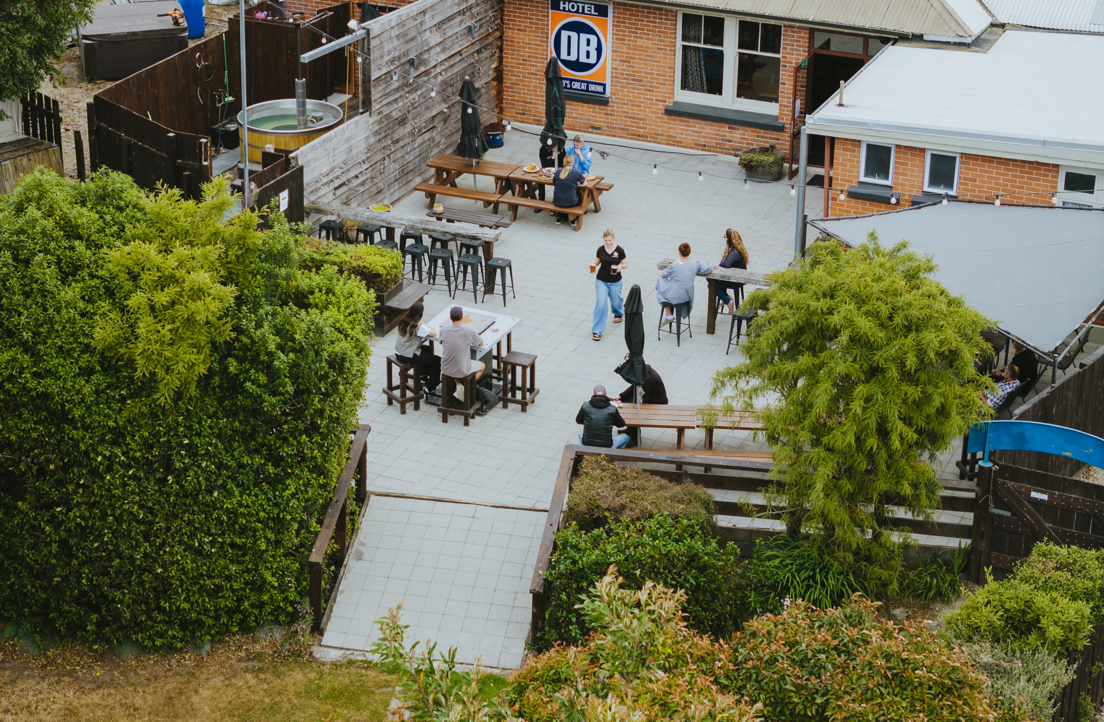 Outdoor patio at a hotel with people sitting at wooden tables and benches surrounded by green bushes and trees.