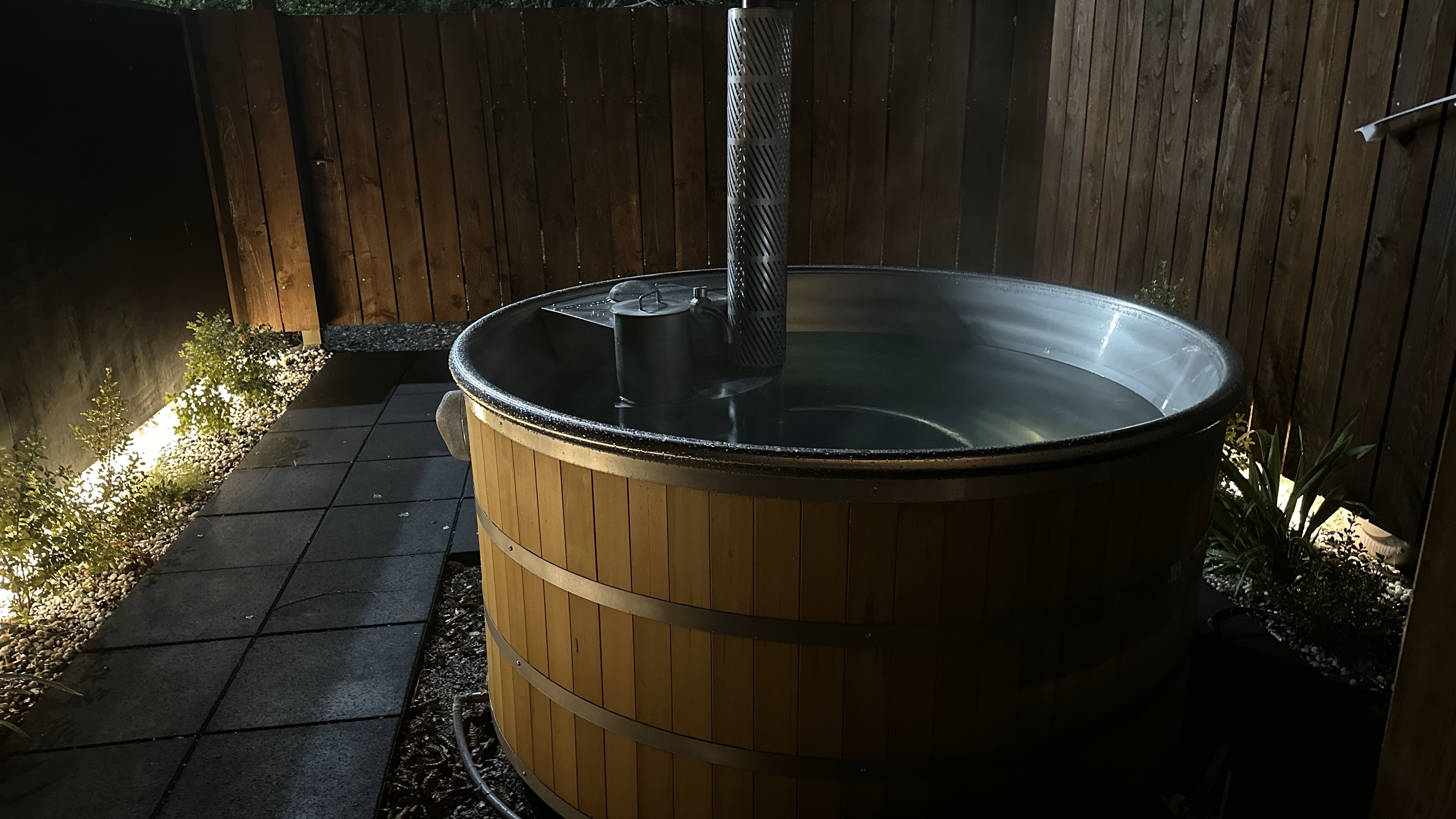 Wooden hot tub filled with water illuminated at night in a fenced backyard.
