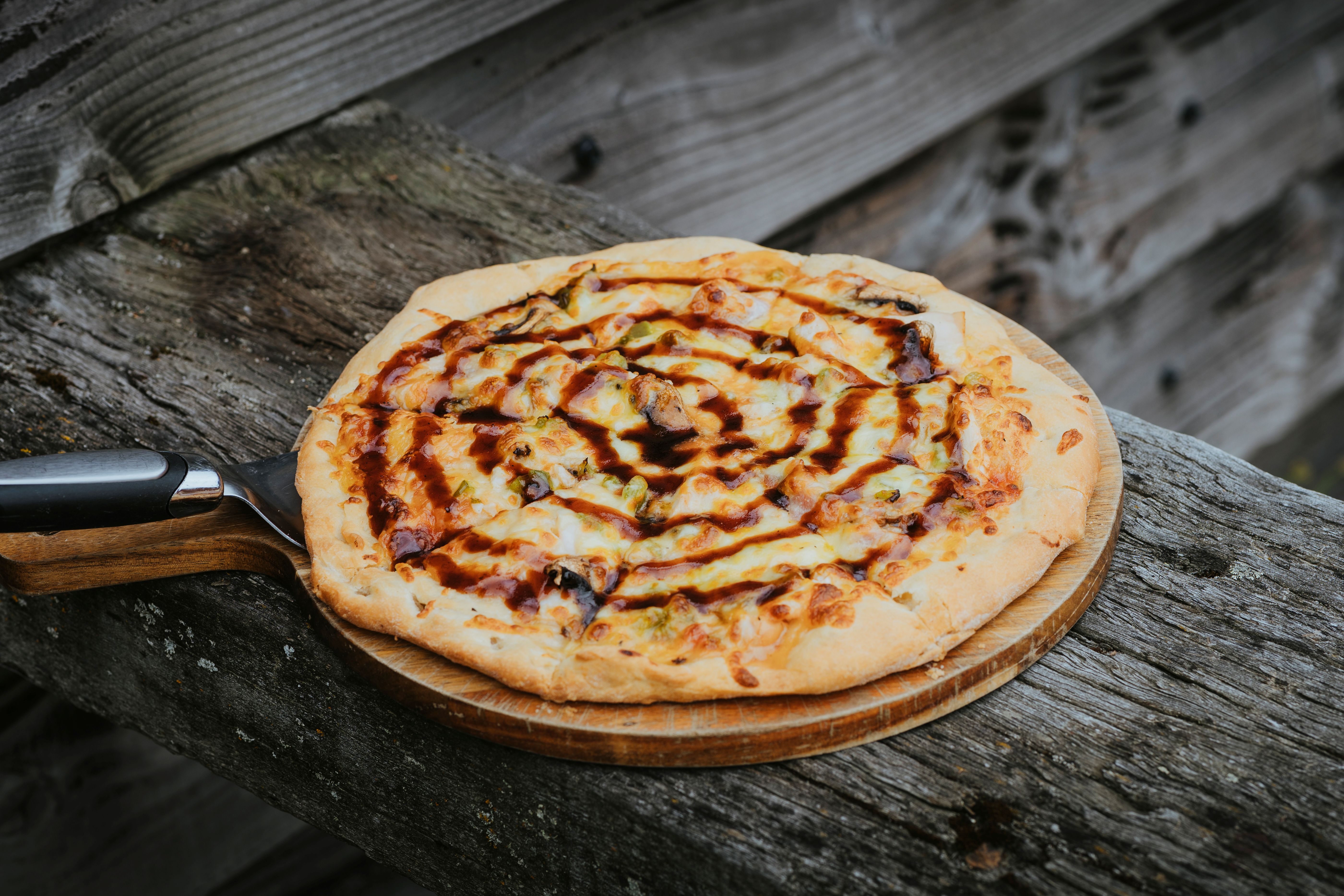 Cheese pizza topped with barbecue sauce drizzle on a wooden pizza board resting on rustic wooden surface.