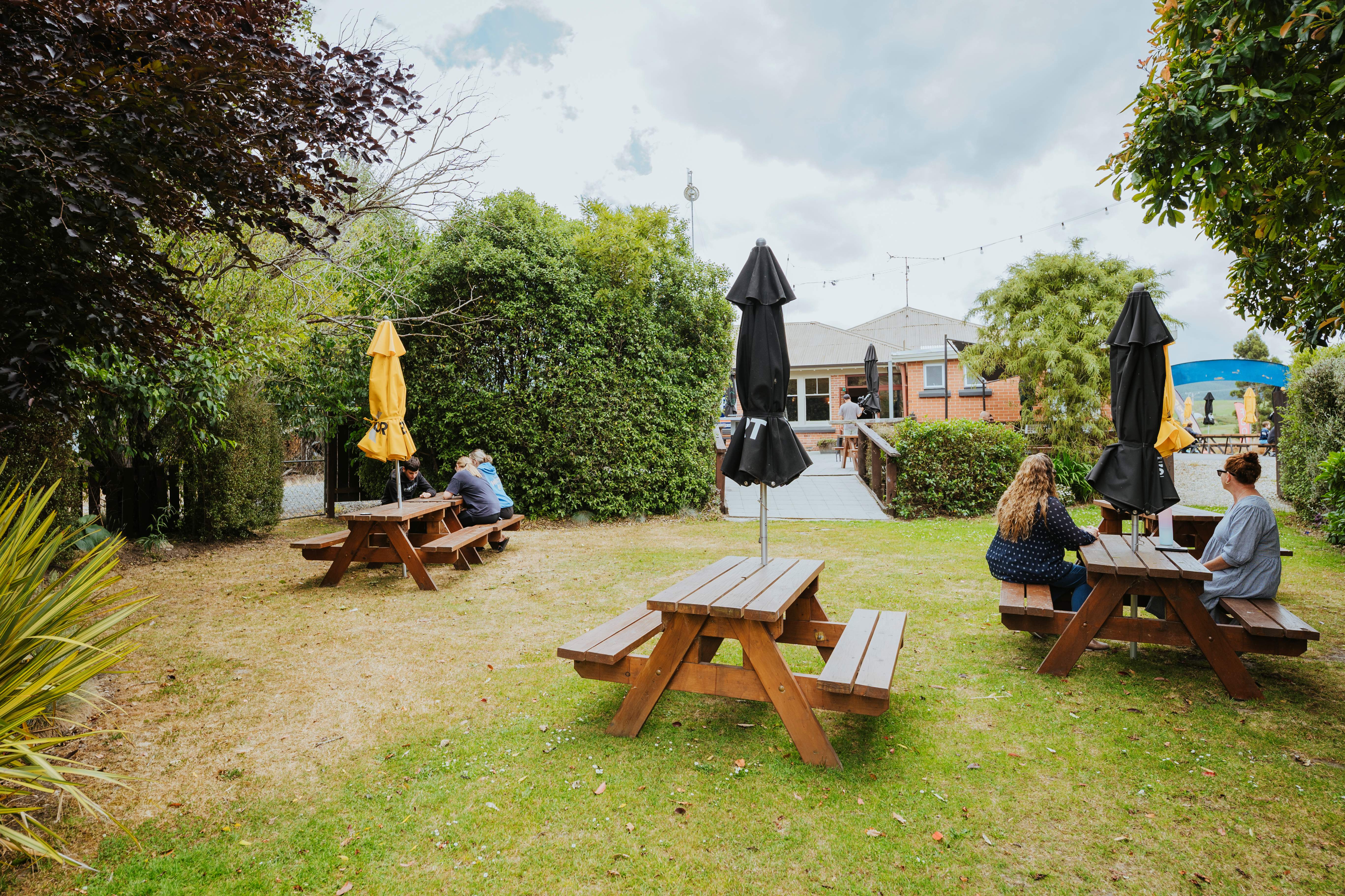 Outdoor beer garden with wooden picnic tables, some with closed umbrellas, and people sitting under trees near a brick building.