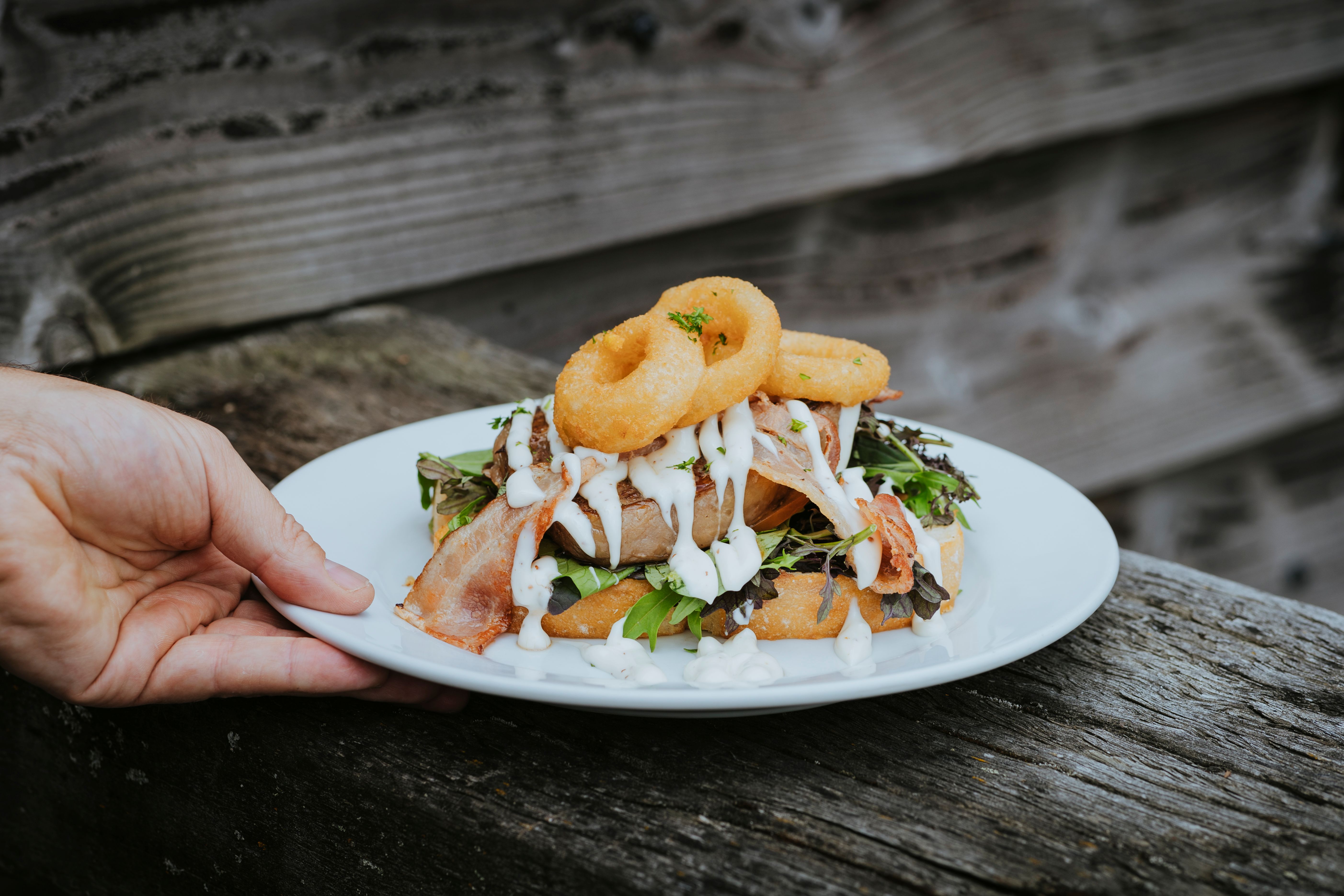 Hand holding a white plate with a gourmet burger topped with onion rings, bacon, lettuce, and white sauce on rustic wooden surface.