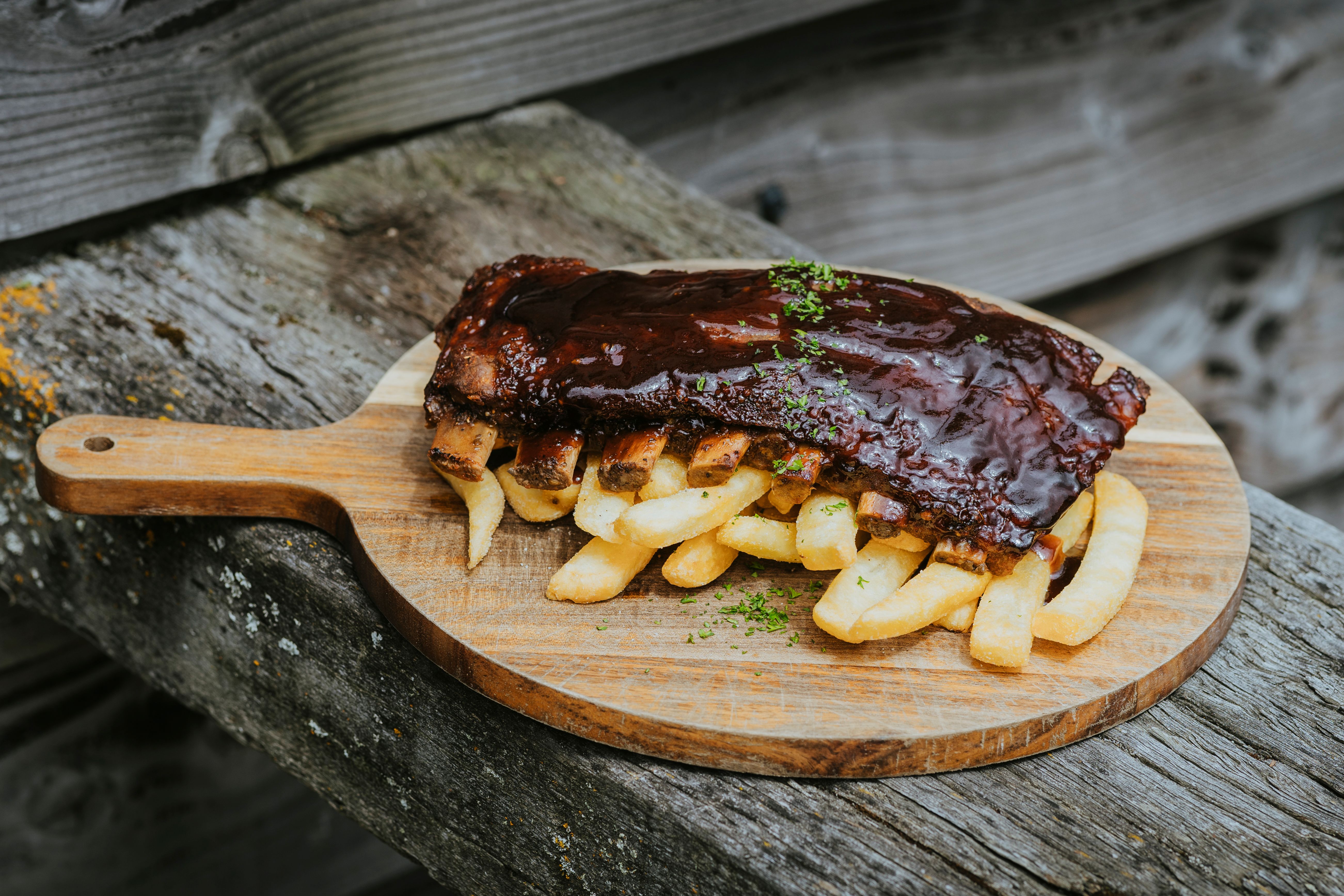 Barbecue pork ribs glazed with sauce served on a bed of thick-cut fries on a wooden paddle board.