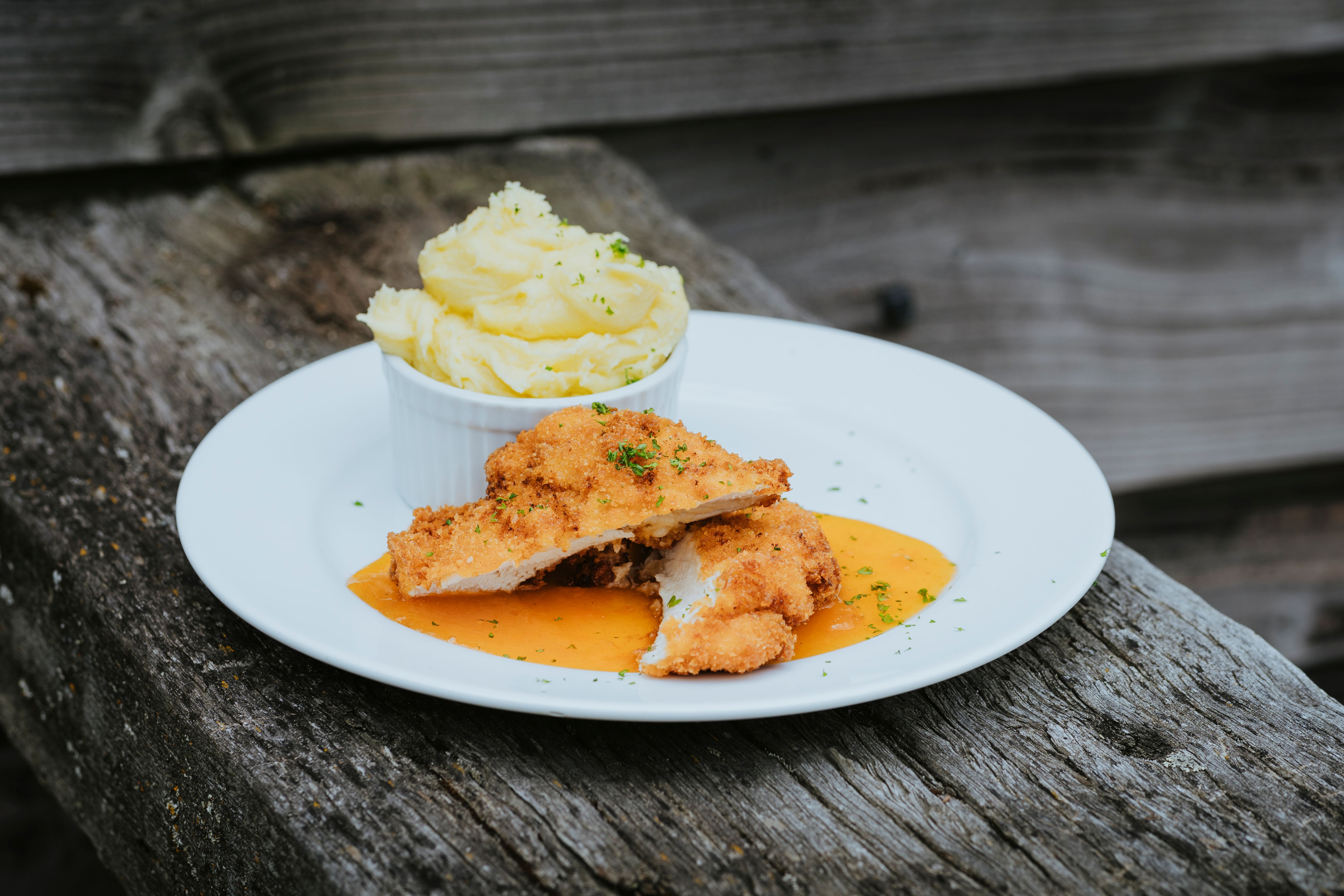 Breaded chicken schnitzel slices served with mashed potatoes in a ramekin and garnished with herbs on a white plate with sauce underneath.