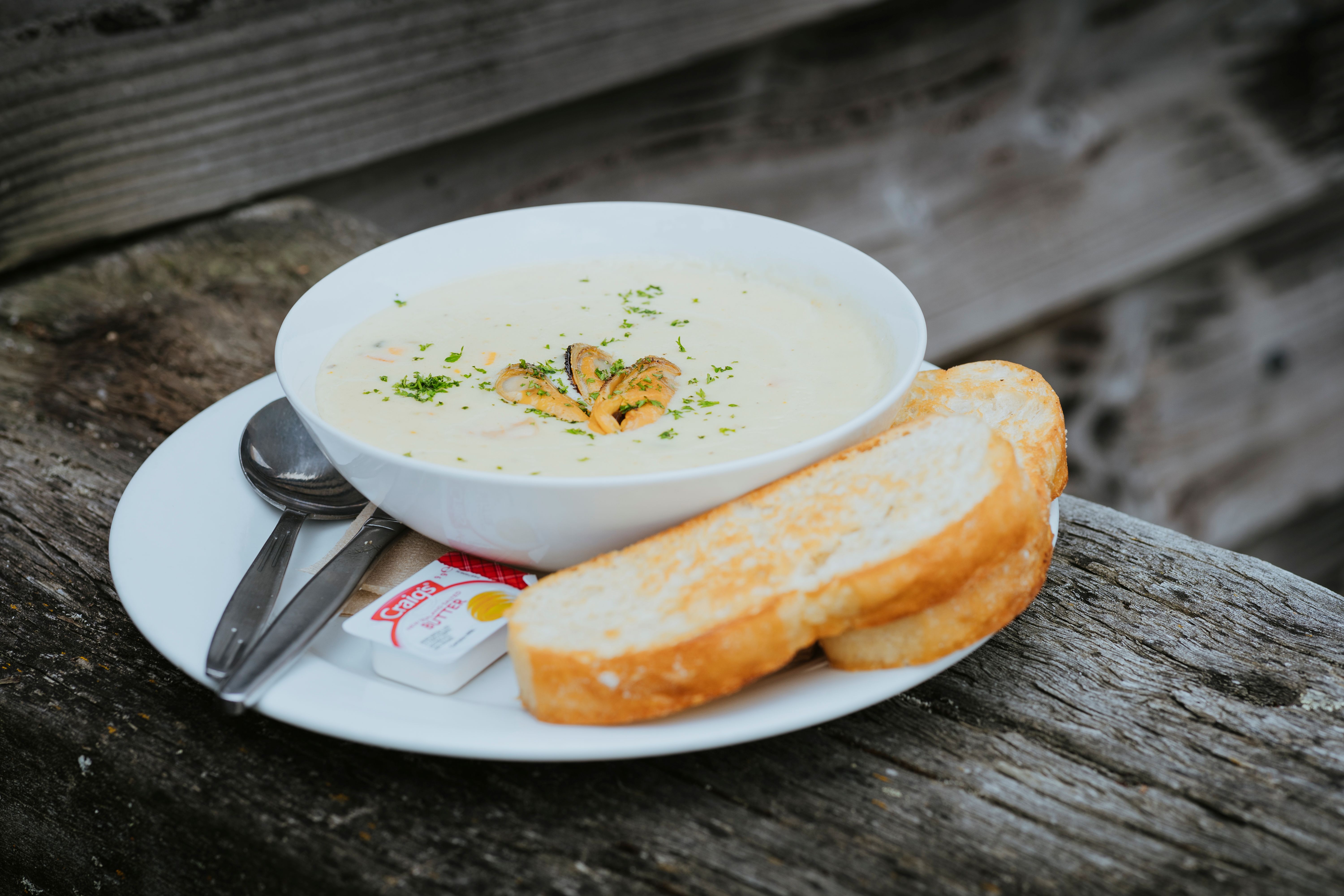 Bowl of creamy seafood chowder garnished with herbs, served with two slices of toasted bread and butter on a white plate with a spoon and fork on a rustic wooden surface.