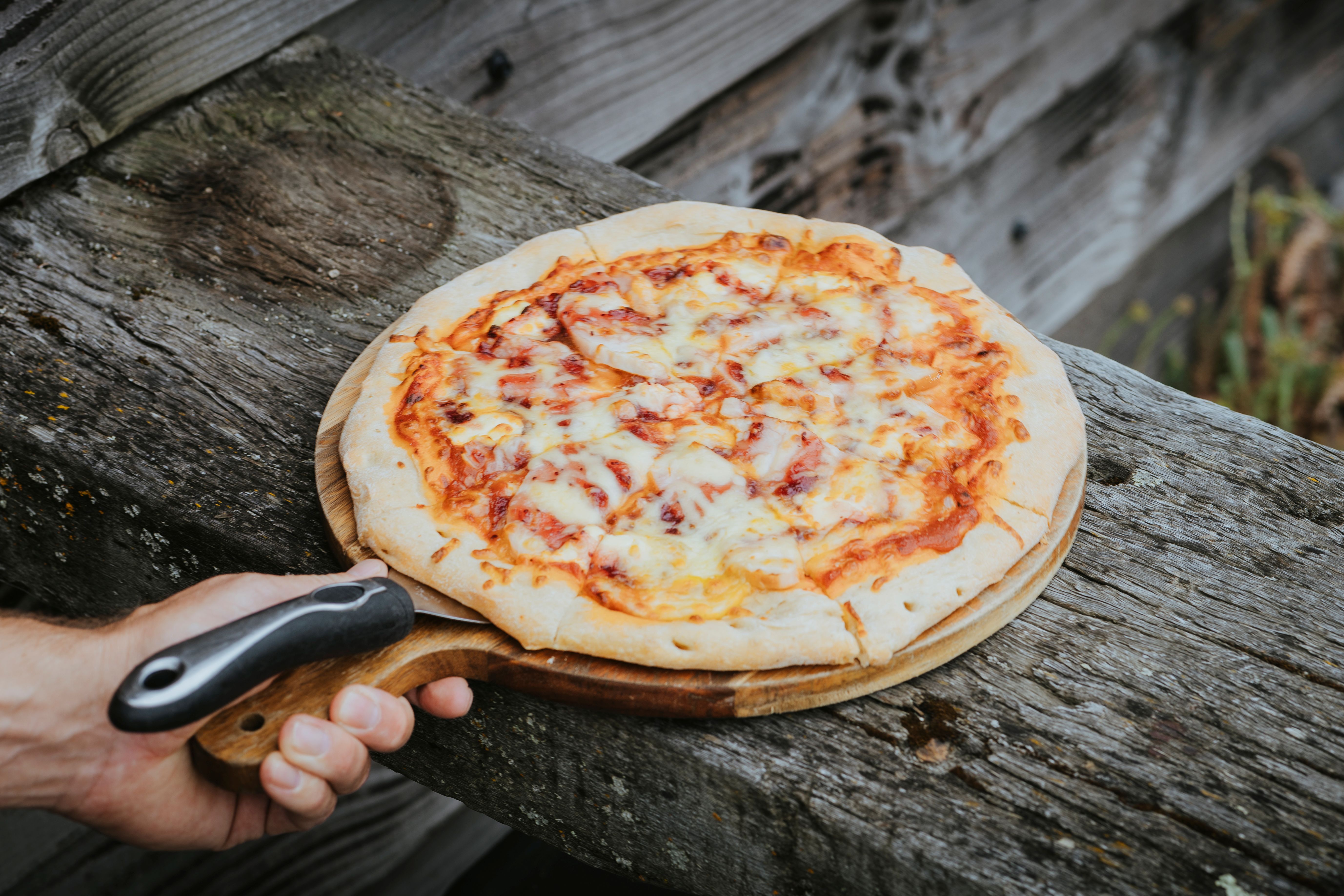 Hand holding a wooden pizza peel with a cheese and tomato pizza on a weathered wooden surface.