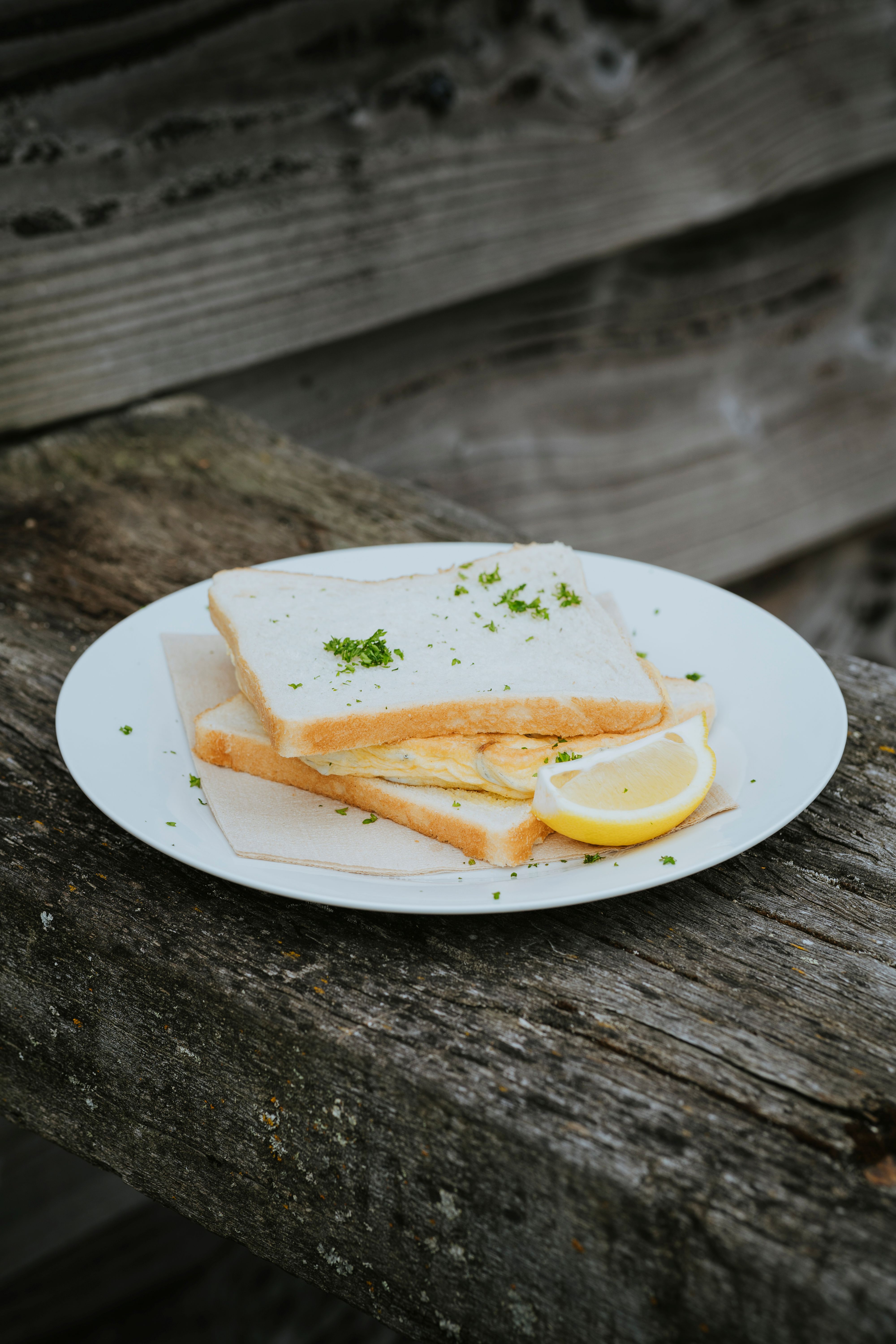 White sandwich with an omelette and parsley garnish served on a white plate with a lemon wedge on a rustic wooden surface.