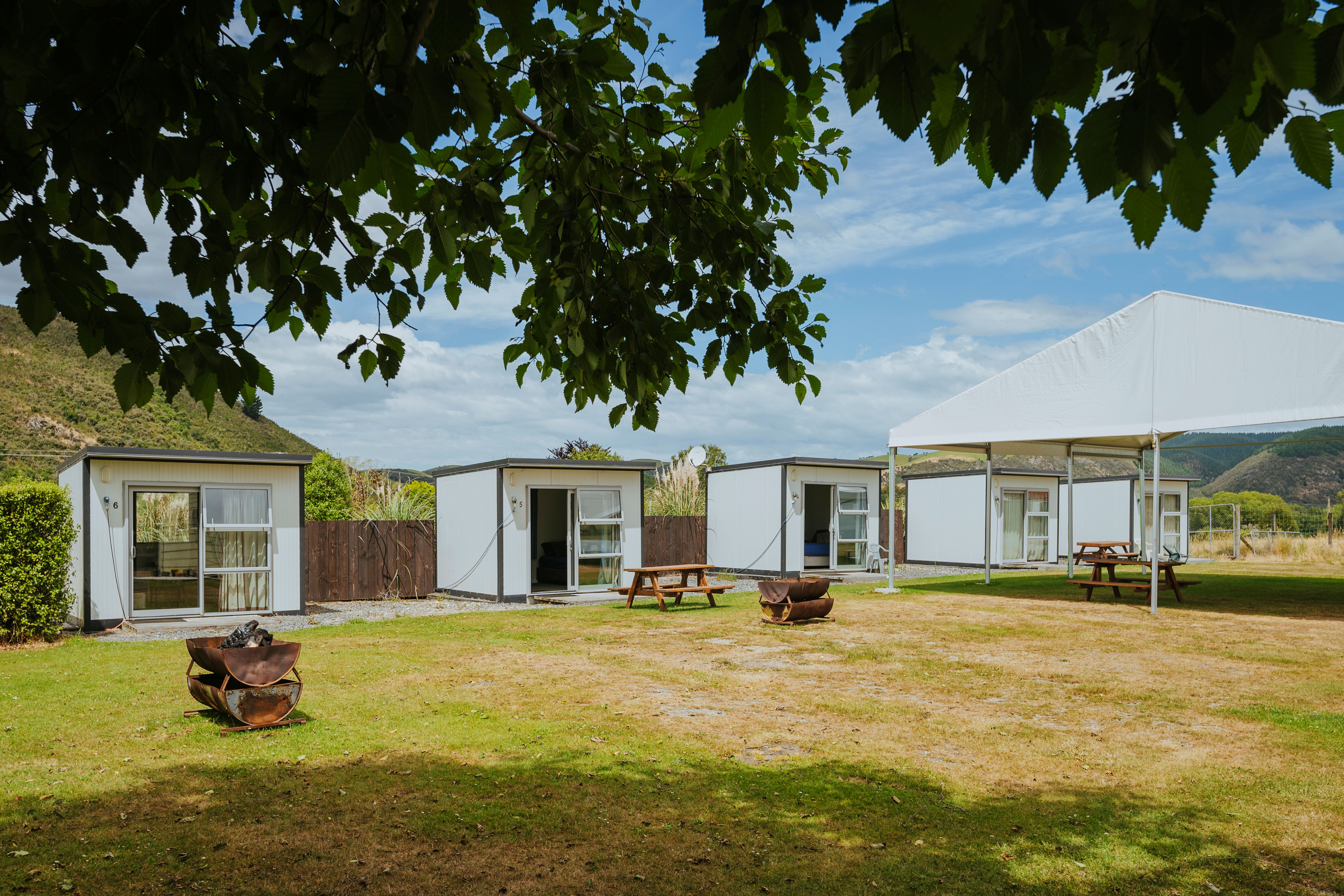 Four small white cabins with sliding glass doors arranged on a grassy area with picnic tables, fire pits, and a white canopy, set against a backdrop of hills and cloudy sky.