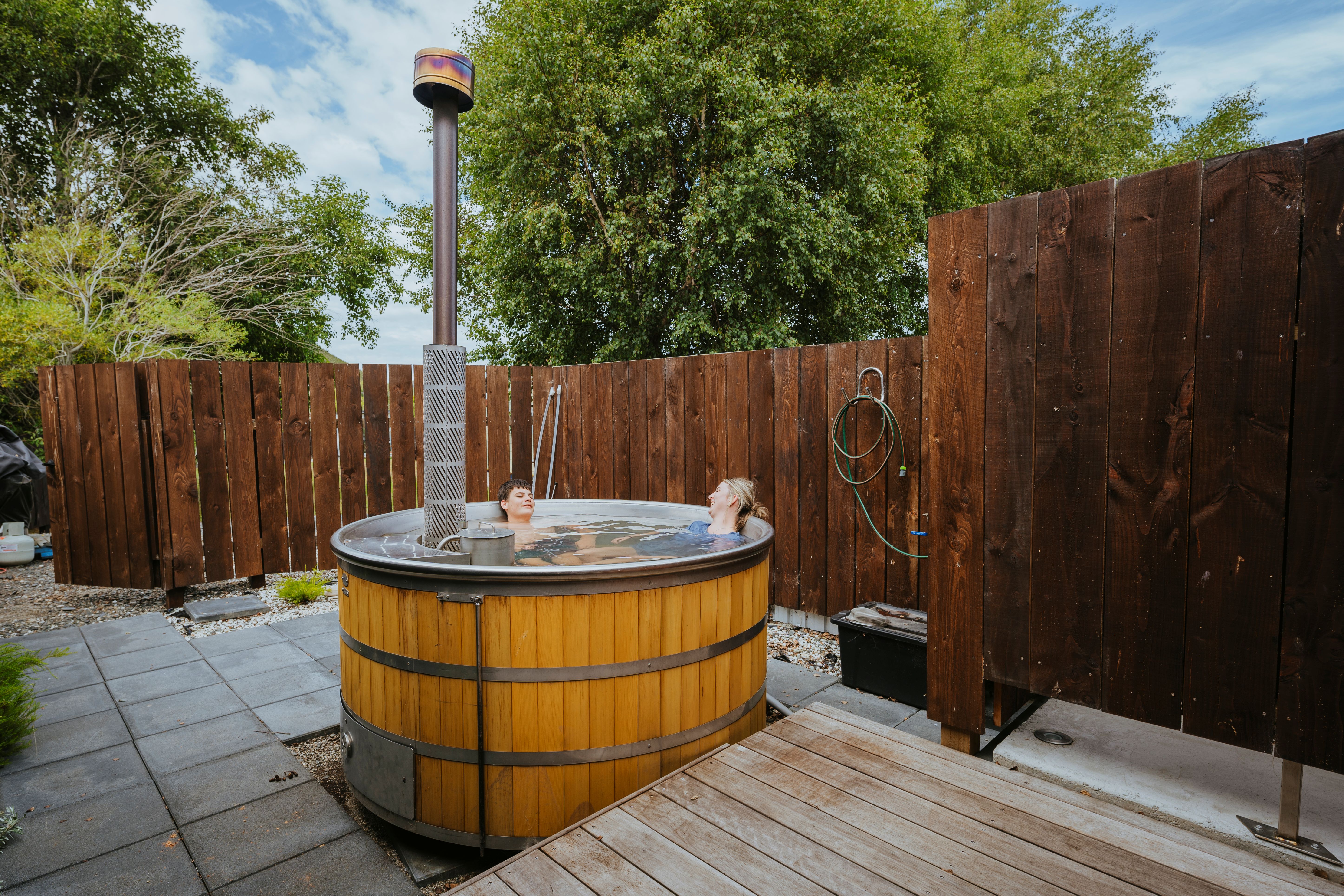 Two people relaxing in a round wooden hot tub outdoors, surrounded by wooden fencing and green trees.