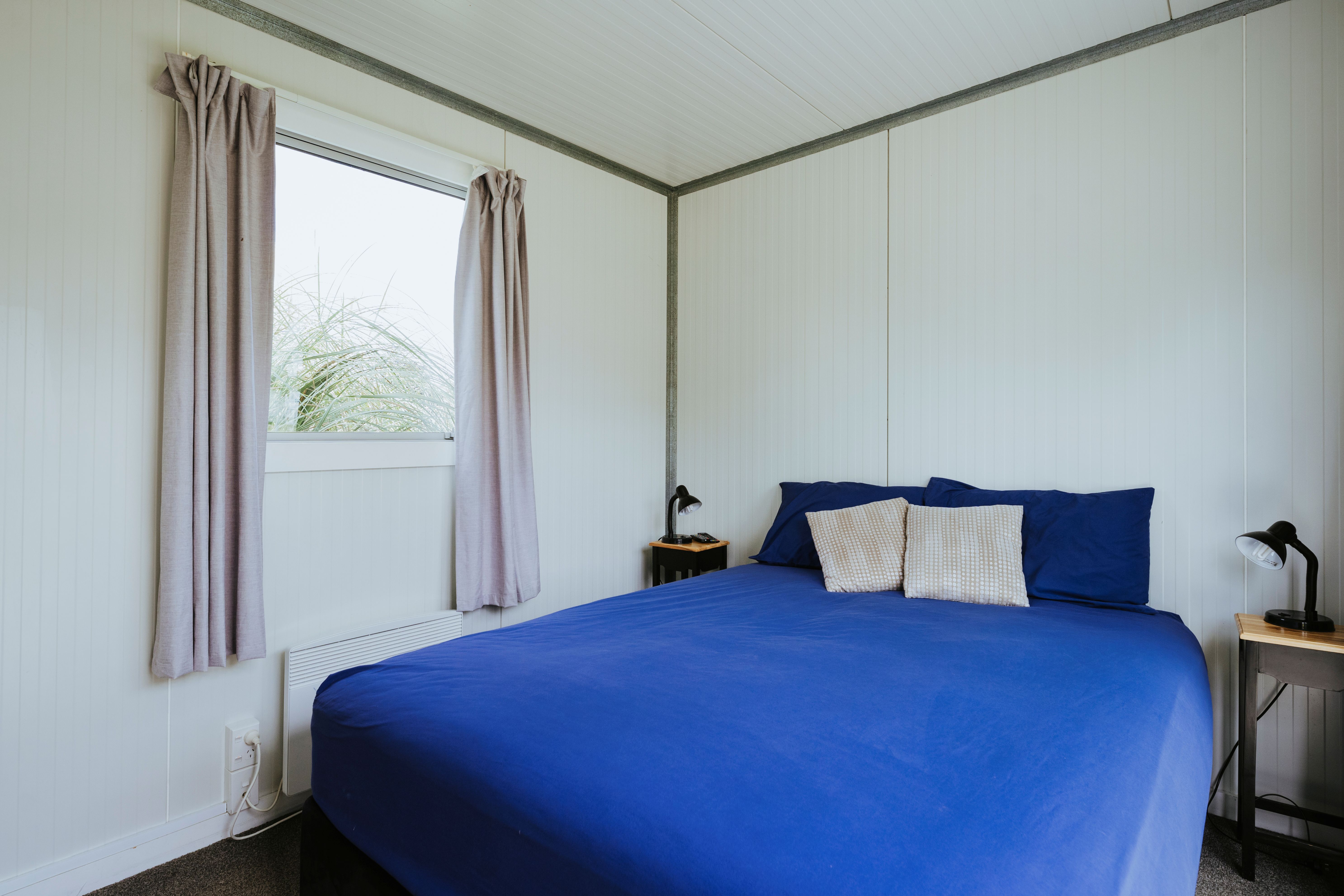 Minimalist bedroom with white paneled walls, a large bed with blue bedding and beige pillows, a window with closed curtains, and two small bedside tables each with a black lamp.