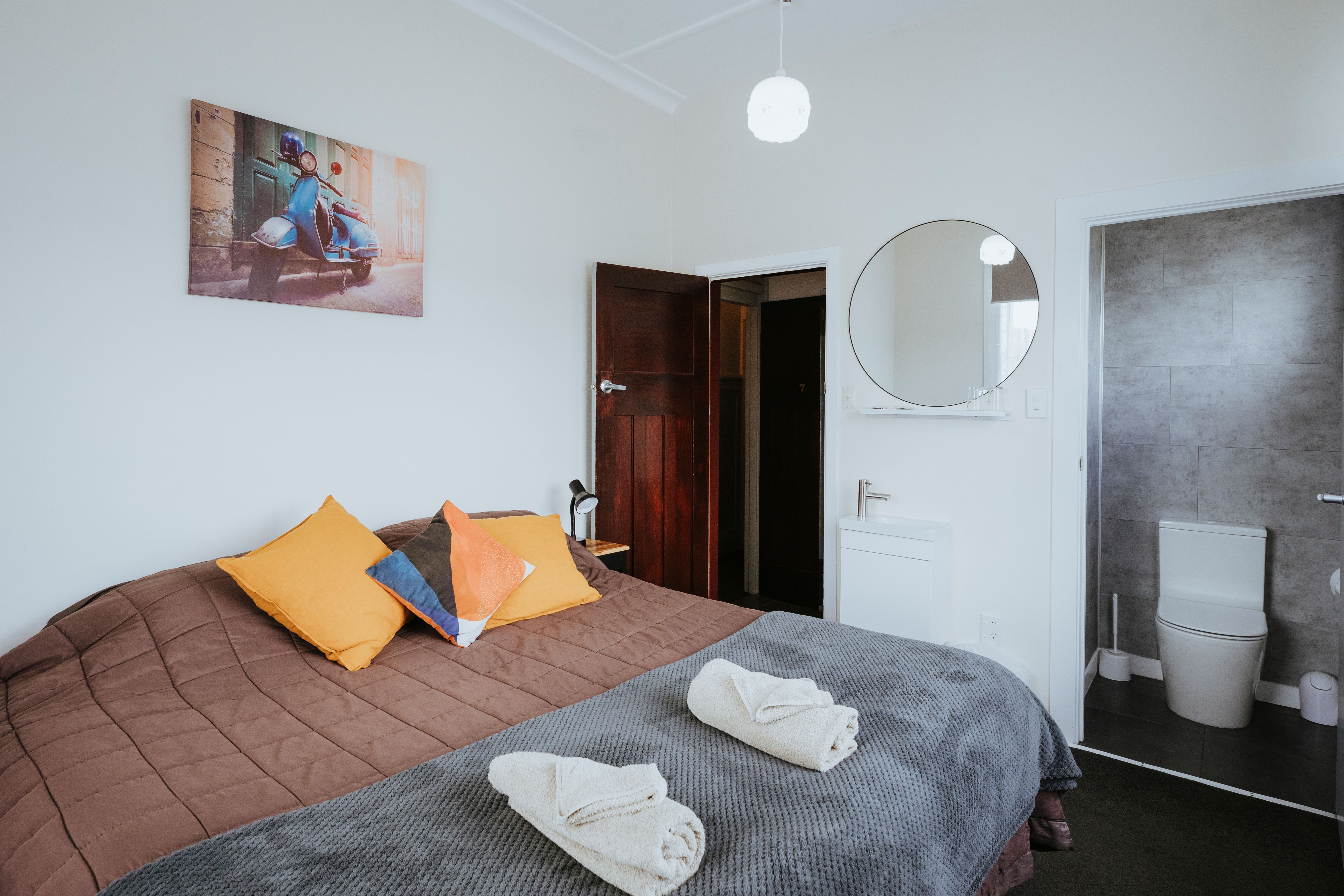 Cozy bedroom with a brown quilt, colorful pillows, two folded white towels, a round mirror, and an adjacent bathroom with a toilet.