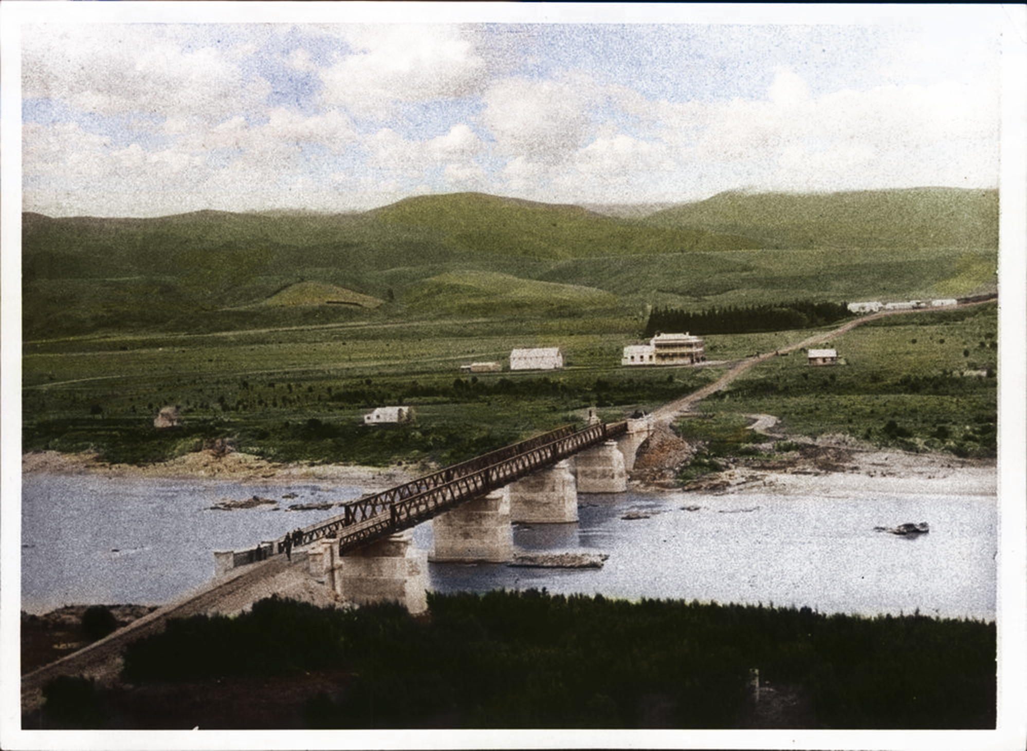 Long wooden bridge with stone pillars crossing a river, leading to a rural area with scattered buildings and green hills under a partly cloudy sky.