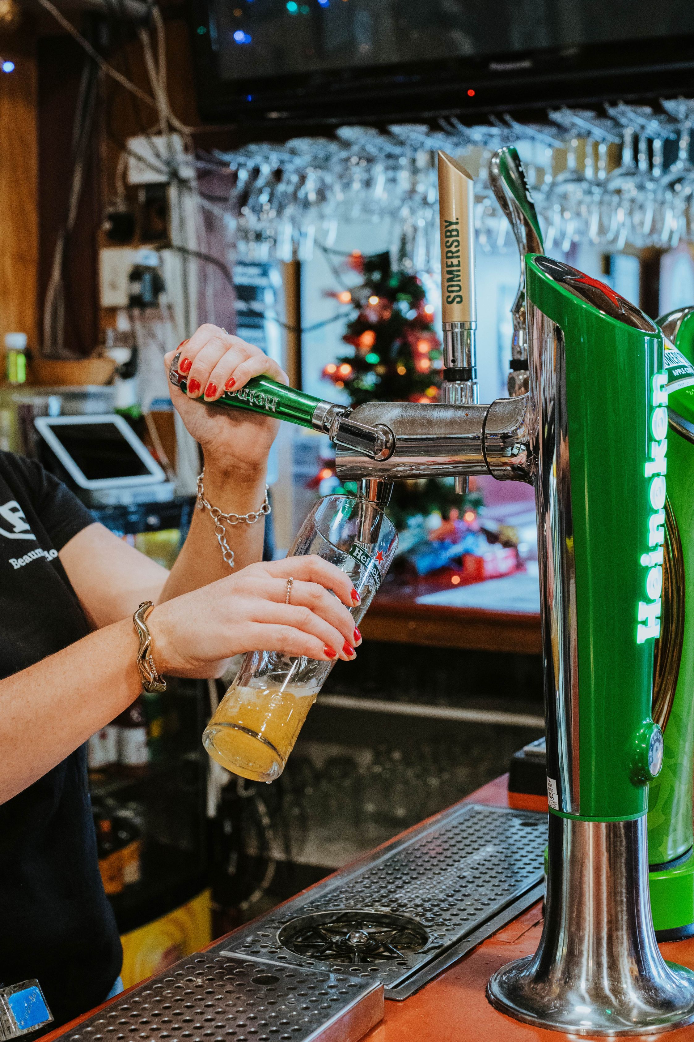 Person with red nail polish pouring beer from a Heineken tap into a glass at a bar with a Christmas tree in the background.