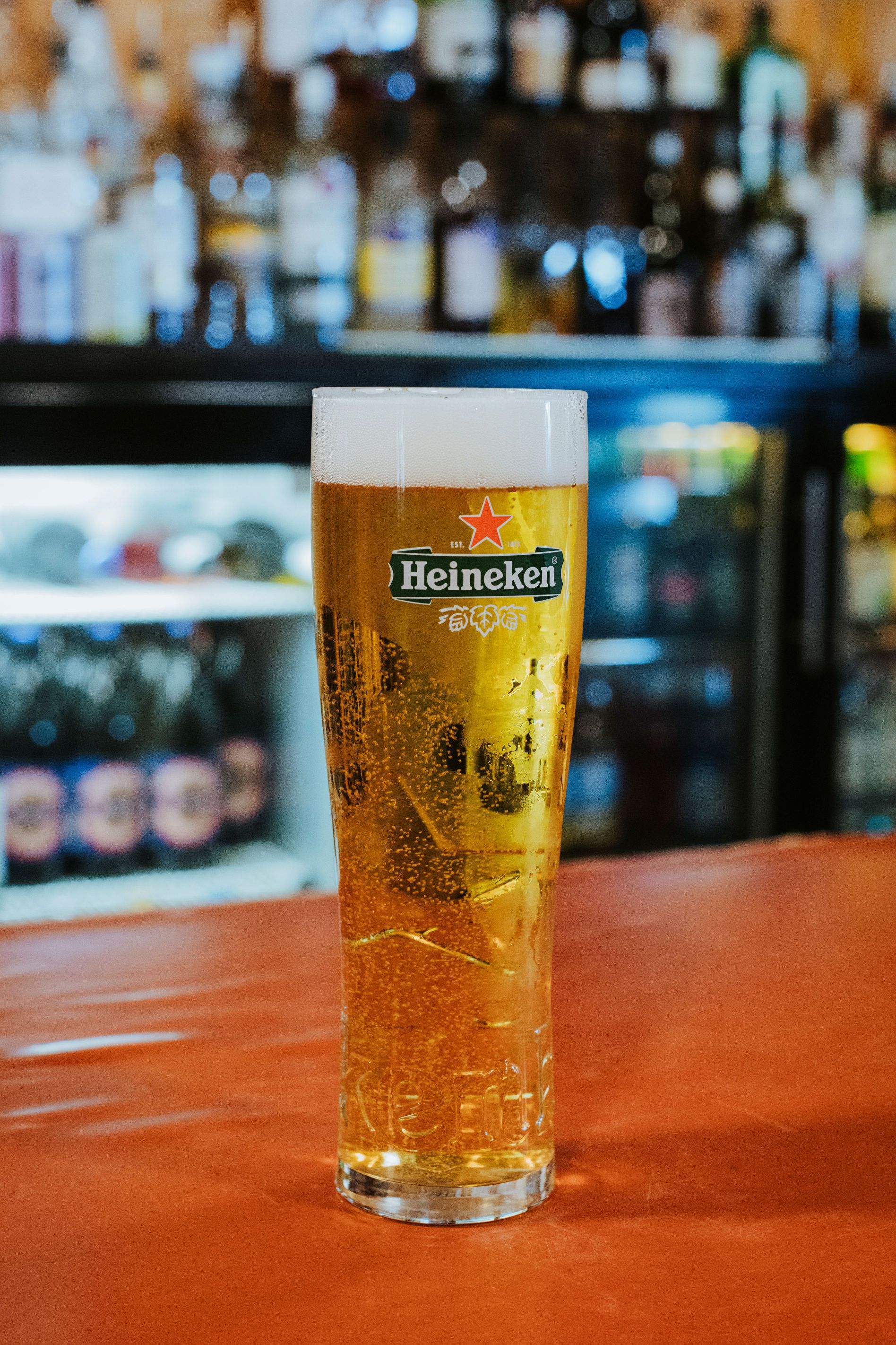 Tall glass of Heineken beer with foam on top, placed on an orange bar counter with blurred bottles in the background.