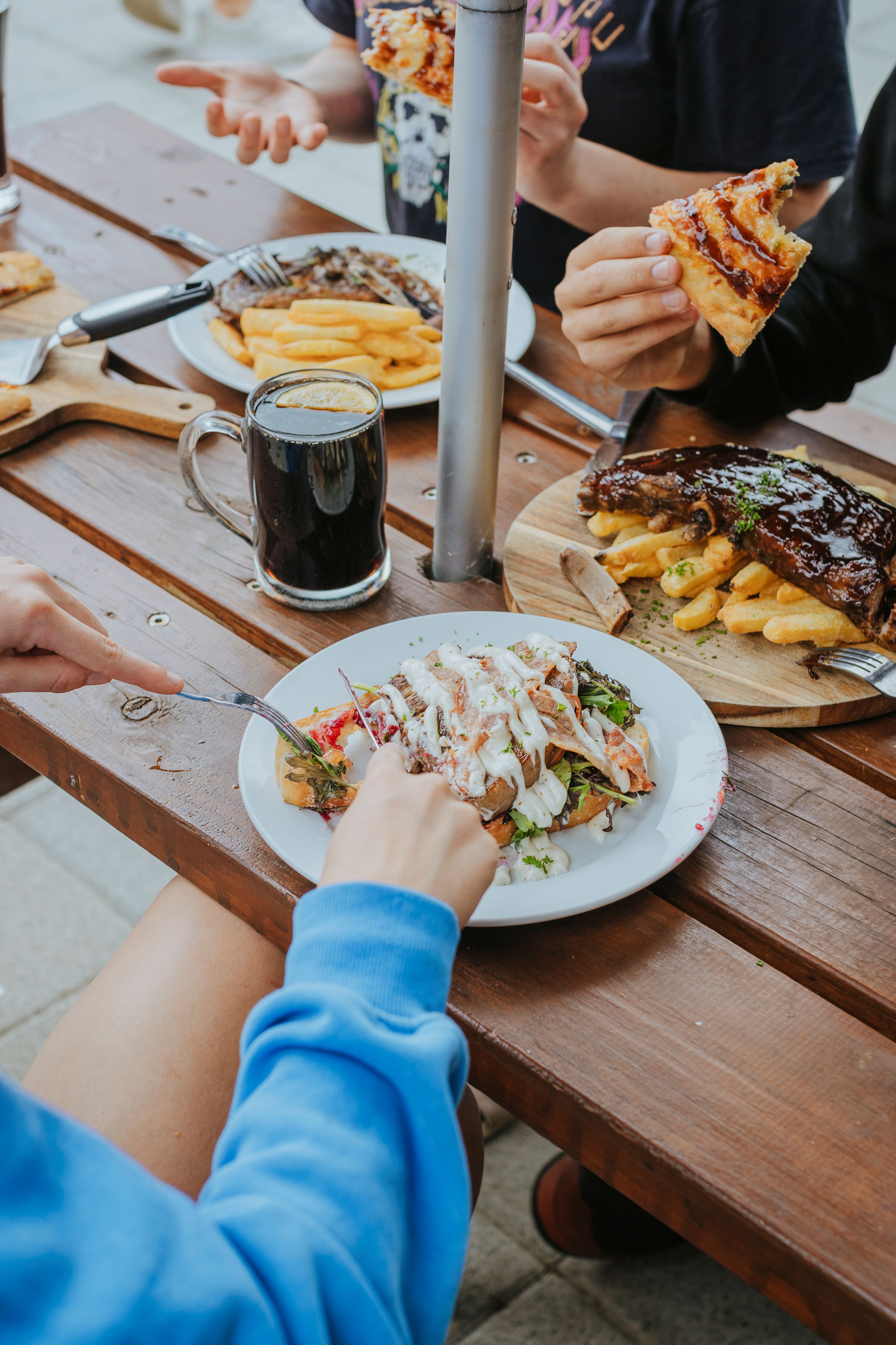 People eating various dishes including ribs with fries, salad with dressing, and pizza around a wooden table outdoors.