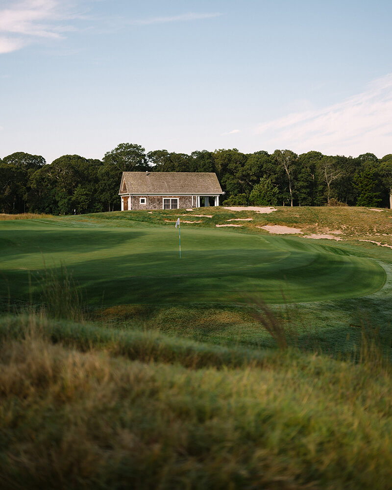 Golf putting green with a flagstick in front of a small wooden clubhouse surrounded by trees under a clear sky.