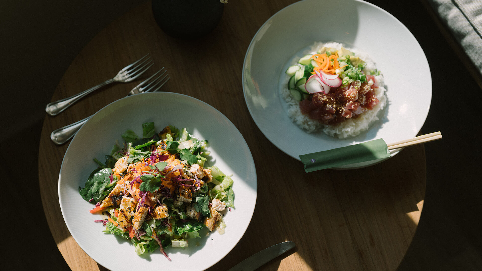 Two plates on a wooden table: a grilled chicken salad with mixed greens and shredded vegetables, and a bowl of rice topped with raw fish, sliced vegetables, and chopsticks wrapped in a green sleeve.