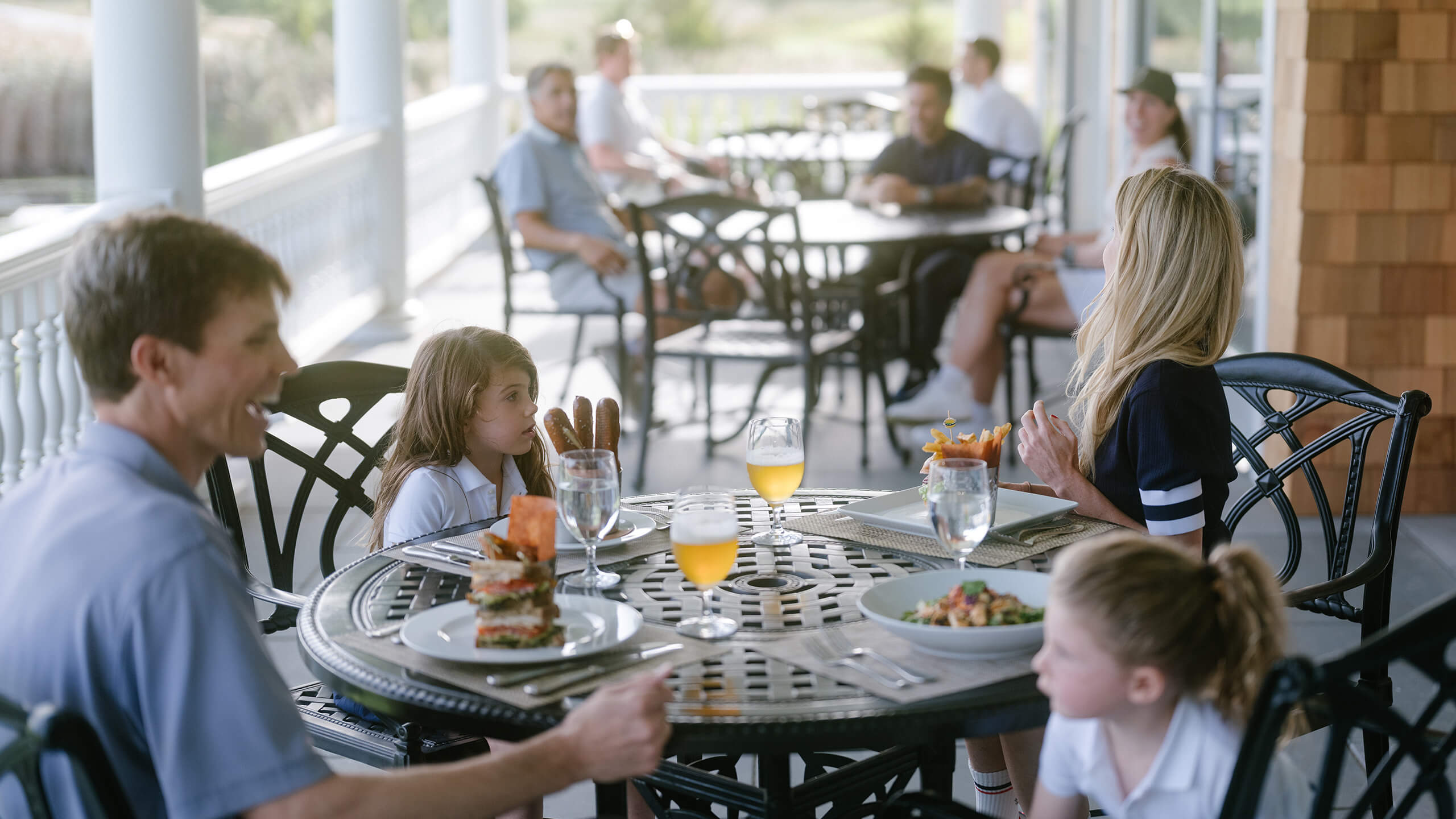 Family dining outdoors on a covered porch at a round metal table with food and drinks during daytime.