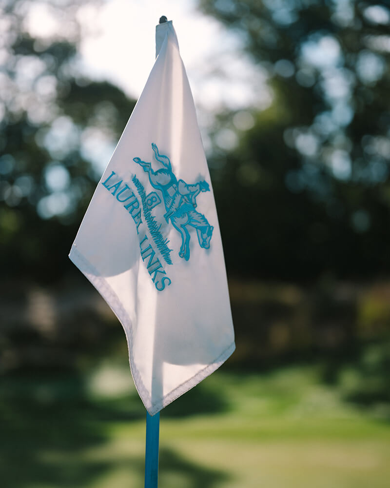 White golf flag with the Laurel Links logo featuring a horse and rider, set against a blurred green golf course background.