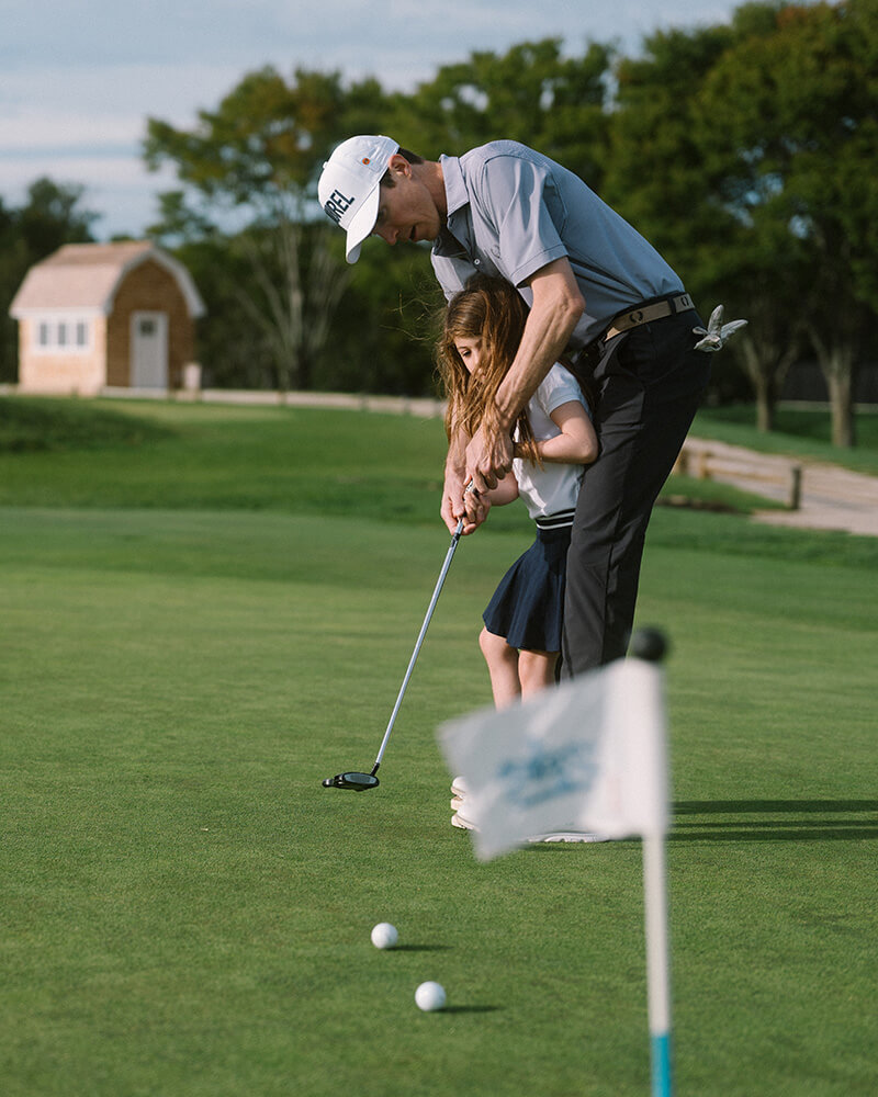 Man teaching a young girl how to putt on a golf green near a hole with a flag.