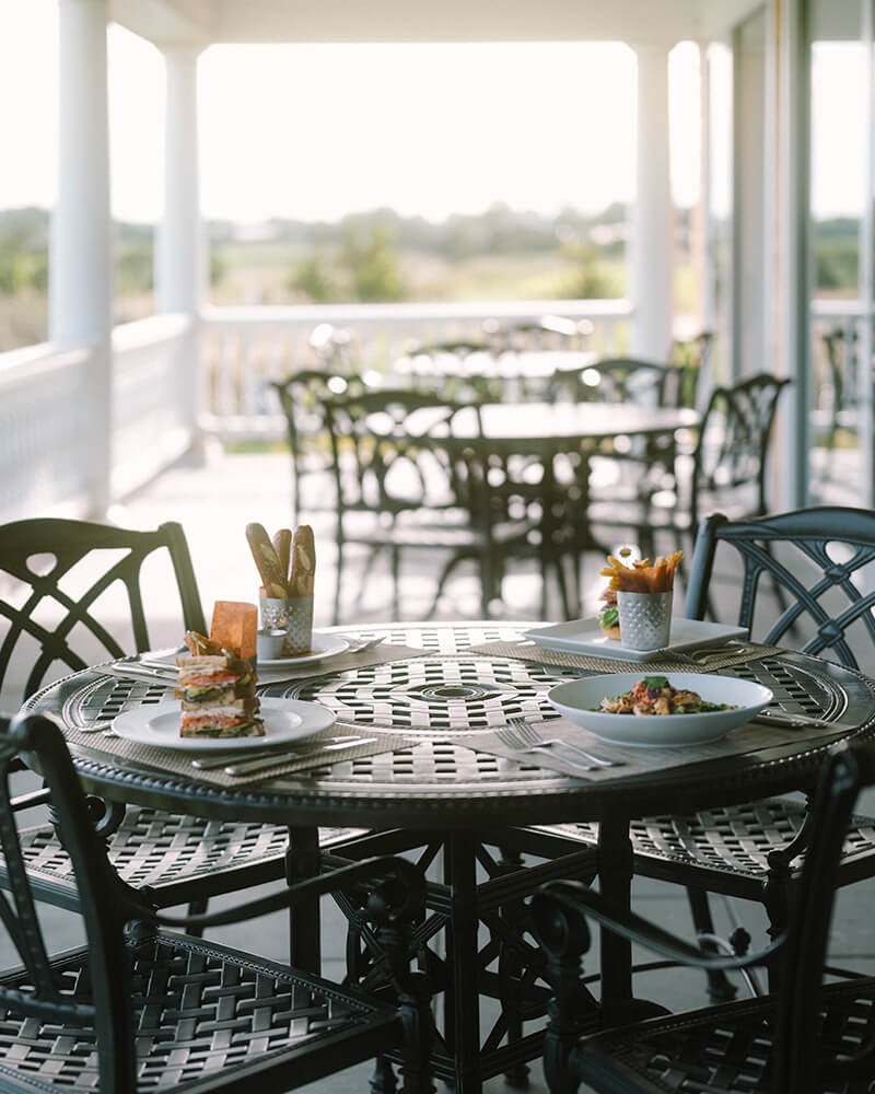Outdoor patio dining area with a round metal table set with plates of food including a sandwich, fries, and a bowl of pasta, surrounded by metal chairs.