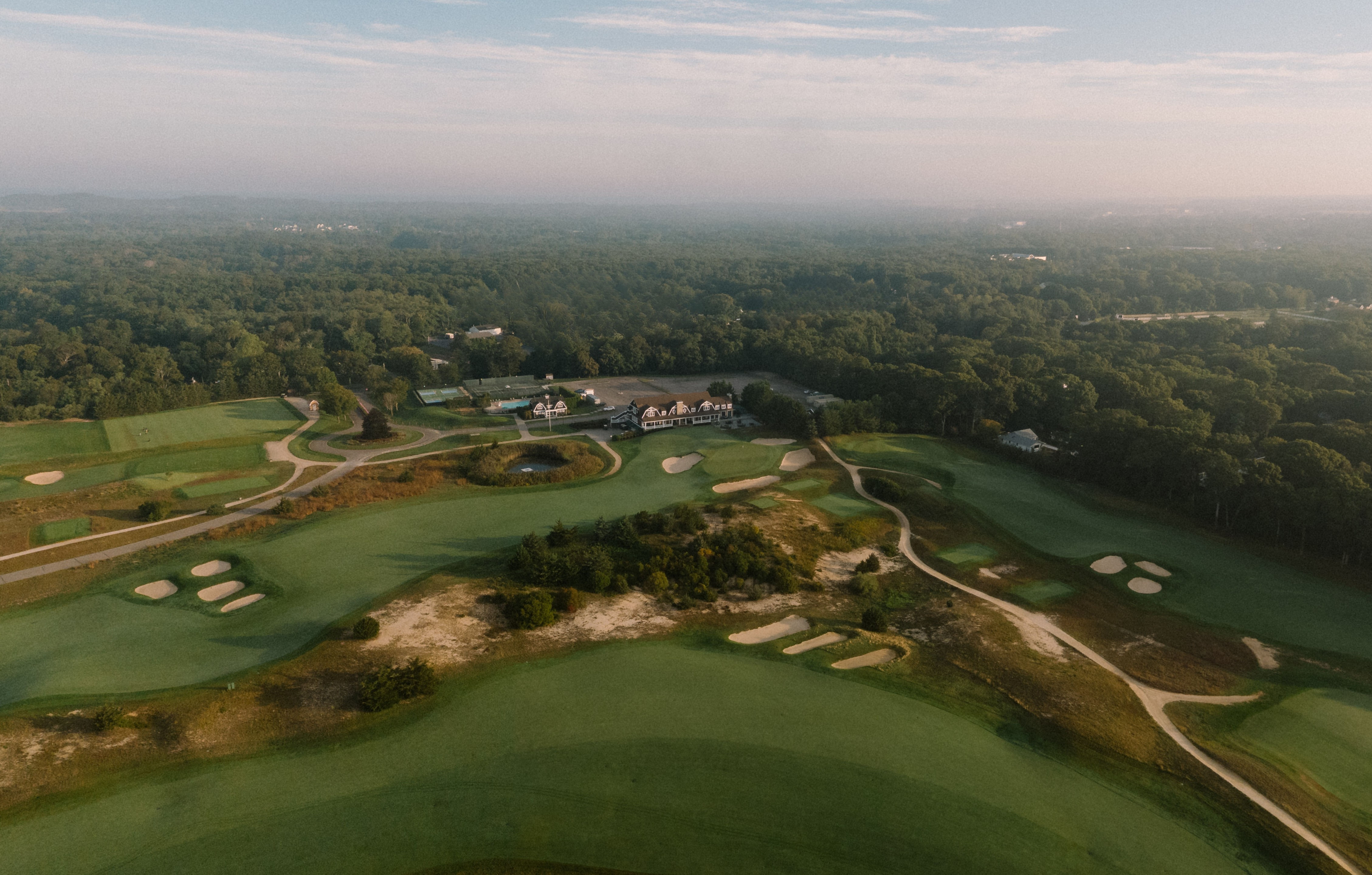 Aerial view of Laurel Links Country Club with green fairways, sand bunkers, a clubhouse, and surrounding dense forest under a partly cloudy sky.