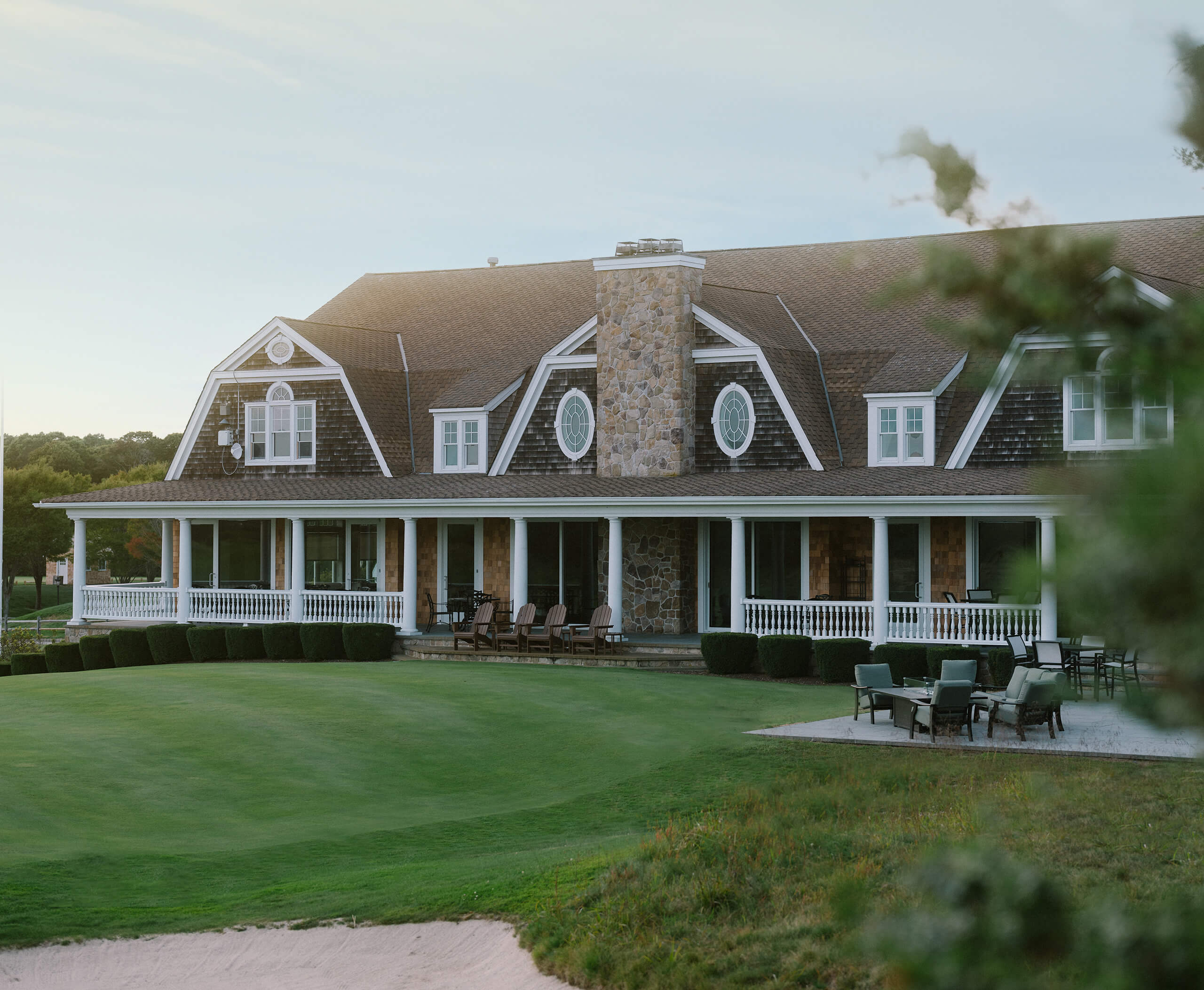 Large rustic clubhouse with a stone chimney, wraparound porch, and outdoor seating overlooking a green golf course with a sand bunker.