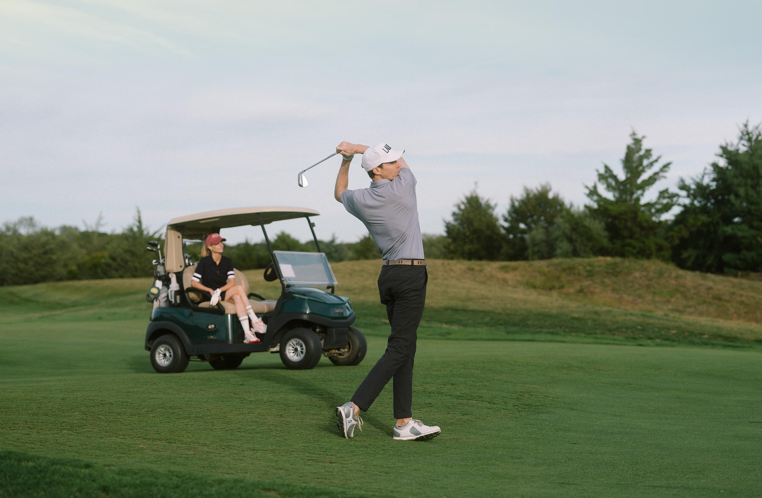Man in gray shirt and white cap swinging a golf club at Laurel Links Country Club with a woman sitting in a nearby golf cart.