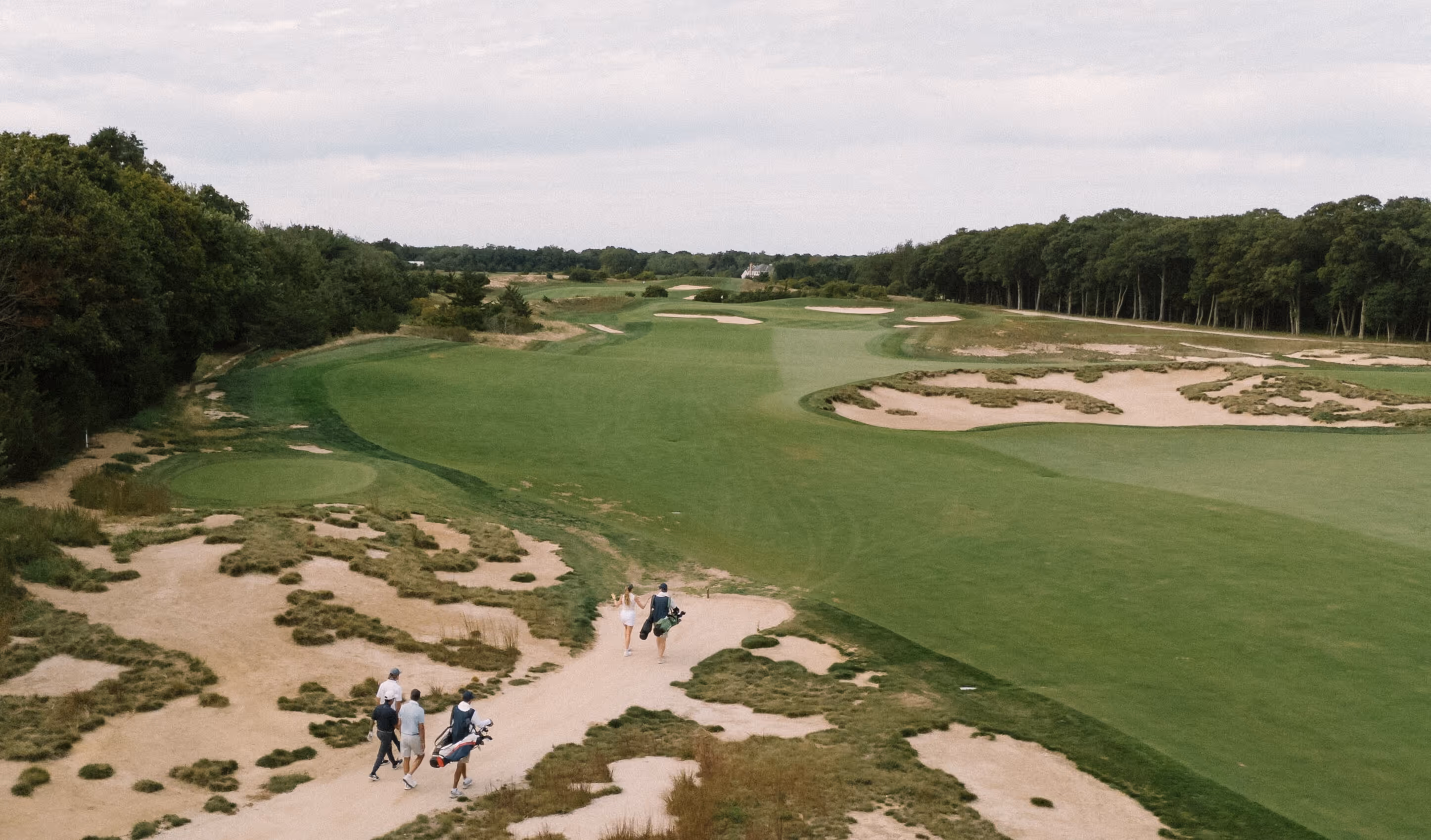 Four golfers walking along a sandy path on Laurel Links Country Club surrounded by green fairways, bunkers, and trees under a cloudy sky.