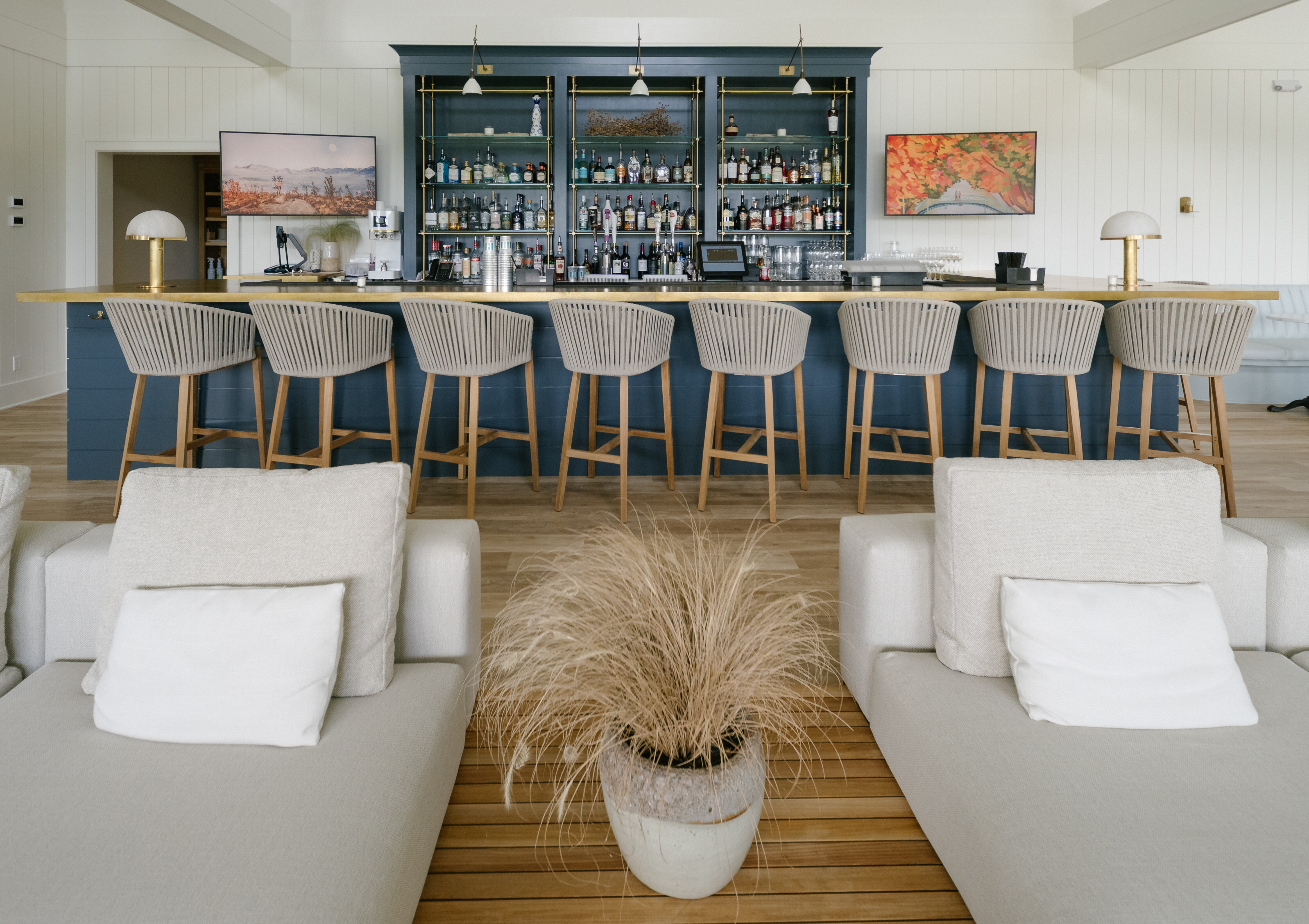 Modern bar with nine wooden stools in front of a blue bar counter and shelves stocked with bottles, viewed from behind light beige cushioned lounge chairs with a potted plant in the center.