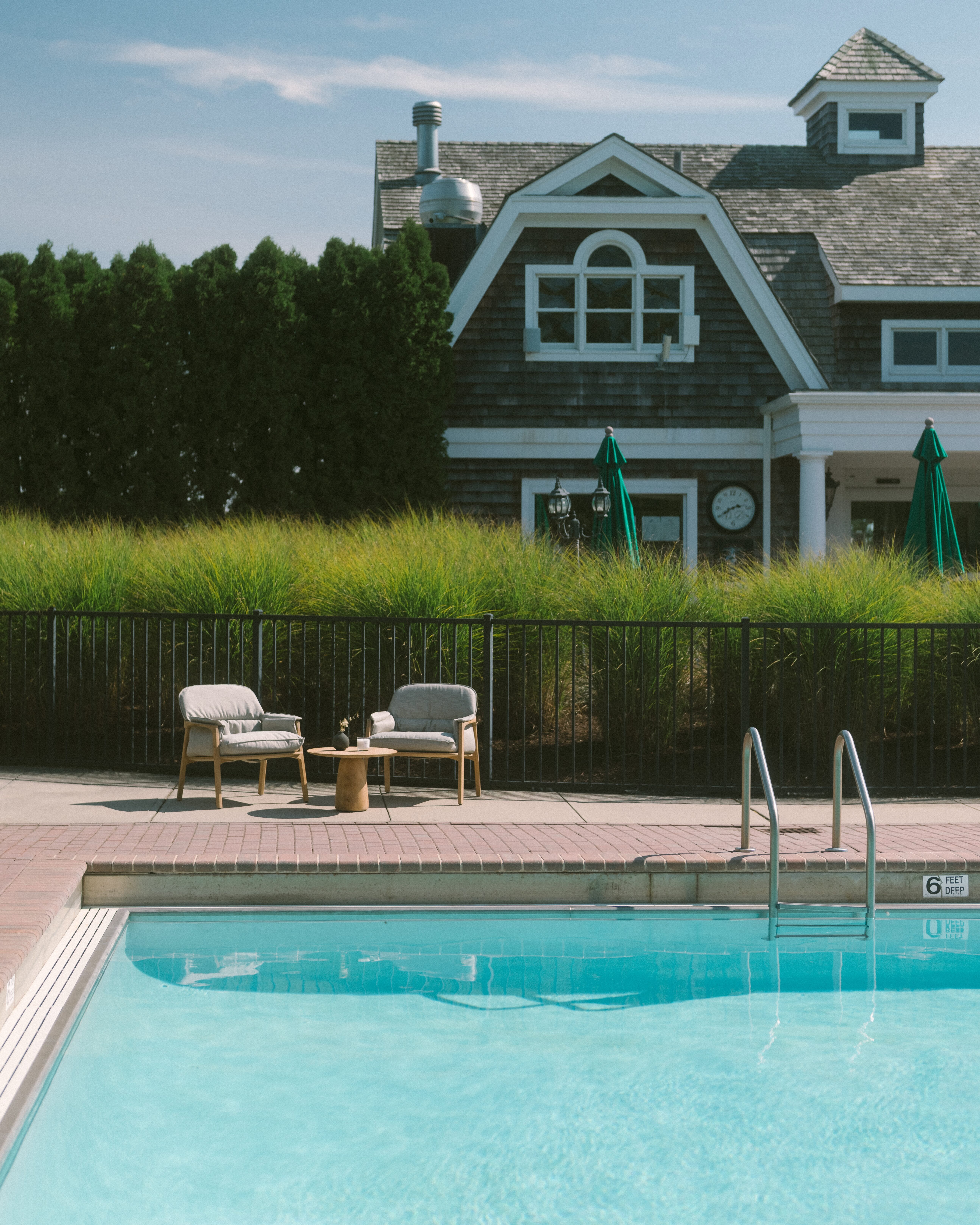 Outdoor pool with metal ladder, two cushioned chairs and a small table on the deck, green shrubs and a house with shingles in the background.