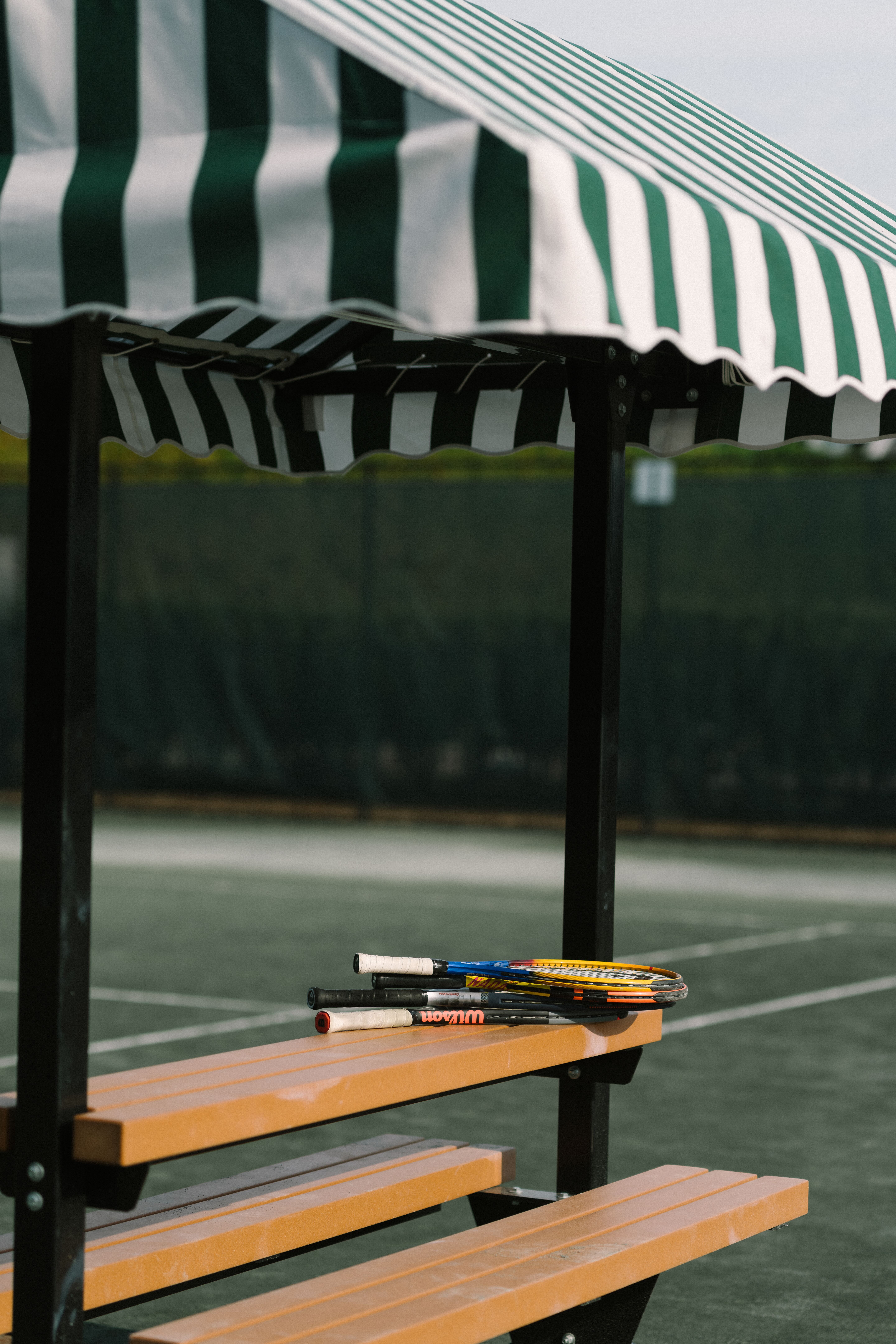 Three tennis rackets resting on a bench under a green and white striped canopy at a tennis court.