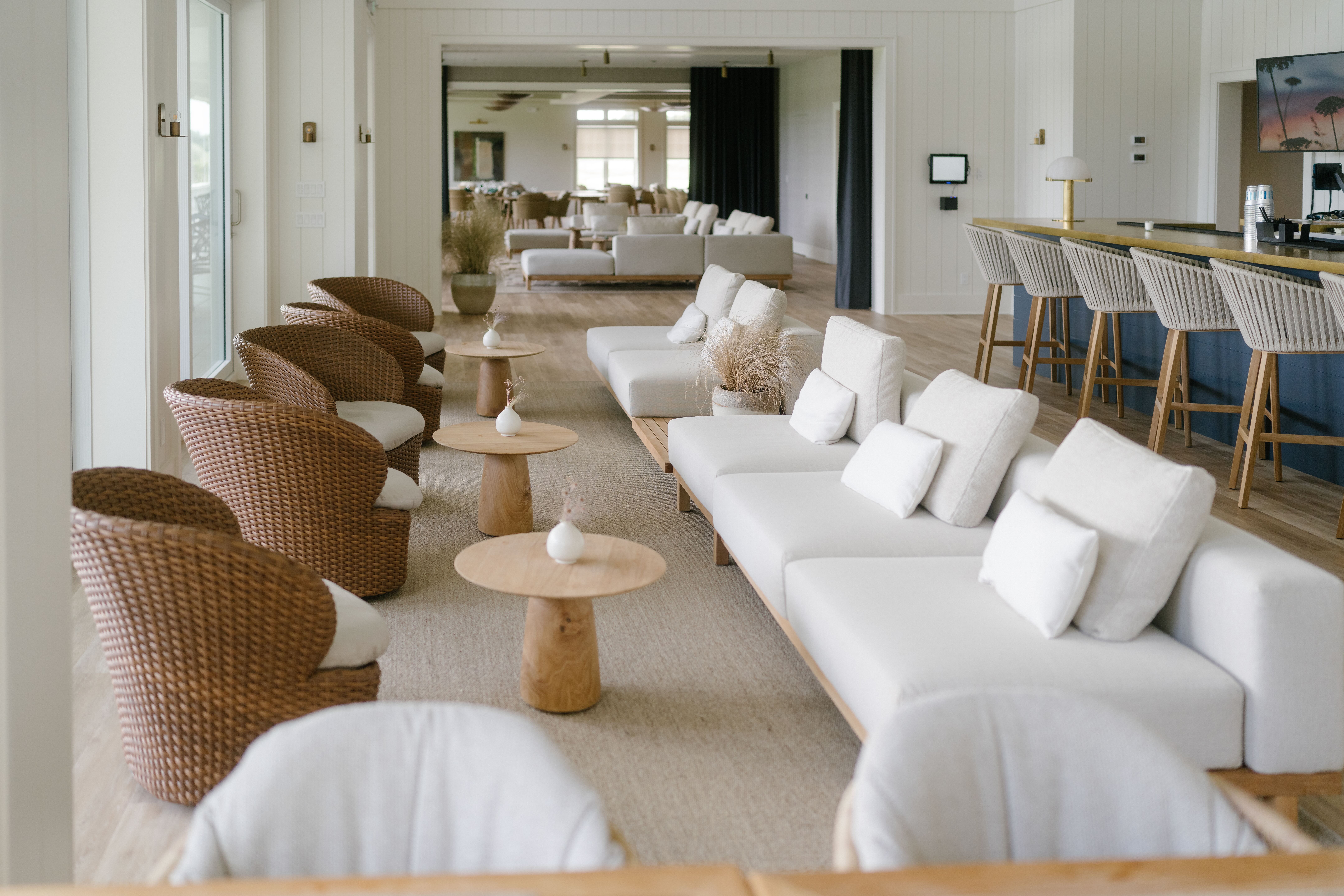 Modern lounge area with wicker armchairs, wooden tables with small white vases, cream sofas, and a bar with high stools in the background.