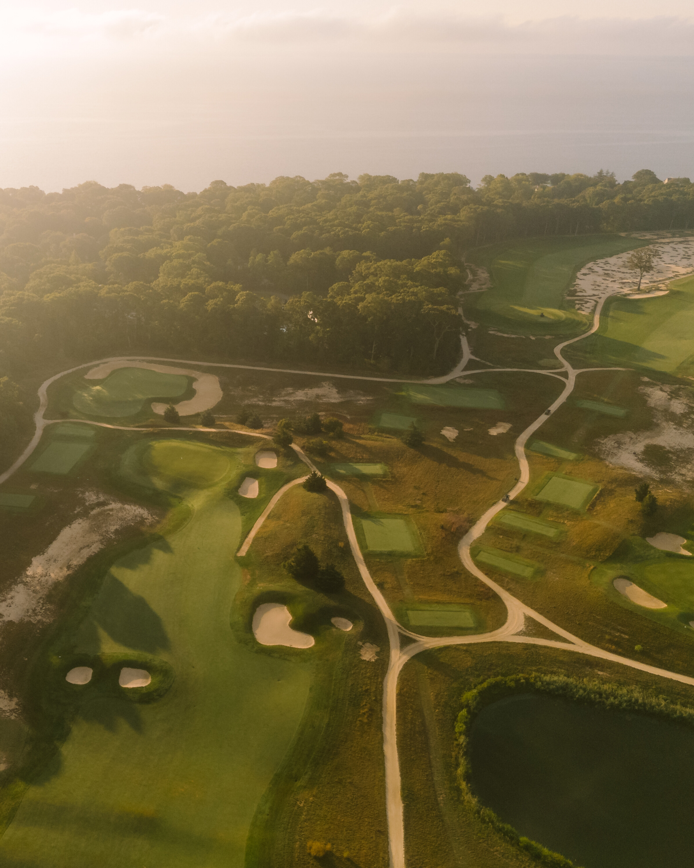 Aerial view of Laurel Links Country Club with sand bunkers, green fairways, winding paths, a pond, and dense forest in the background under soft sunlight.