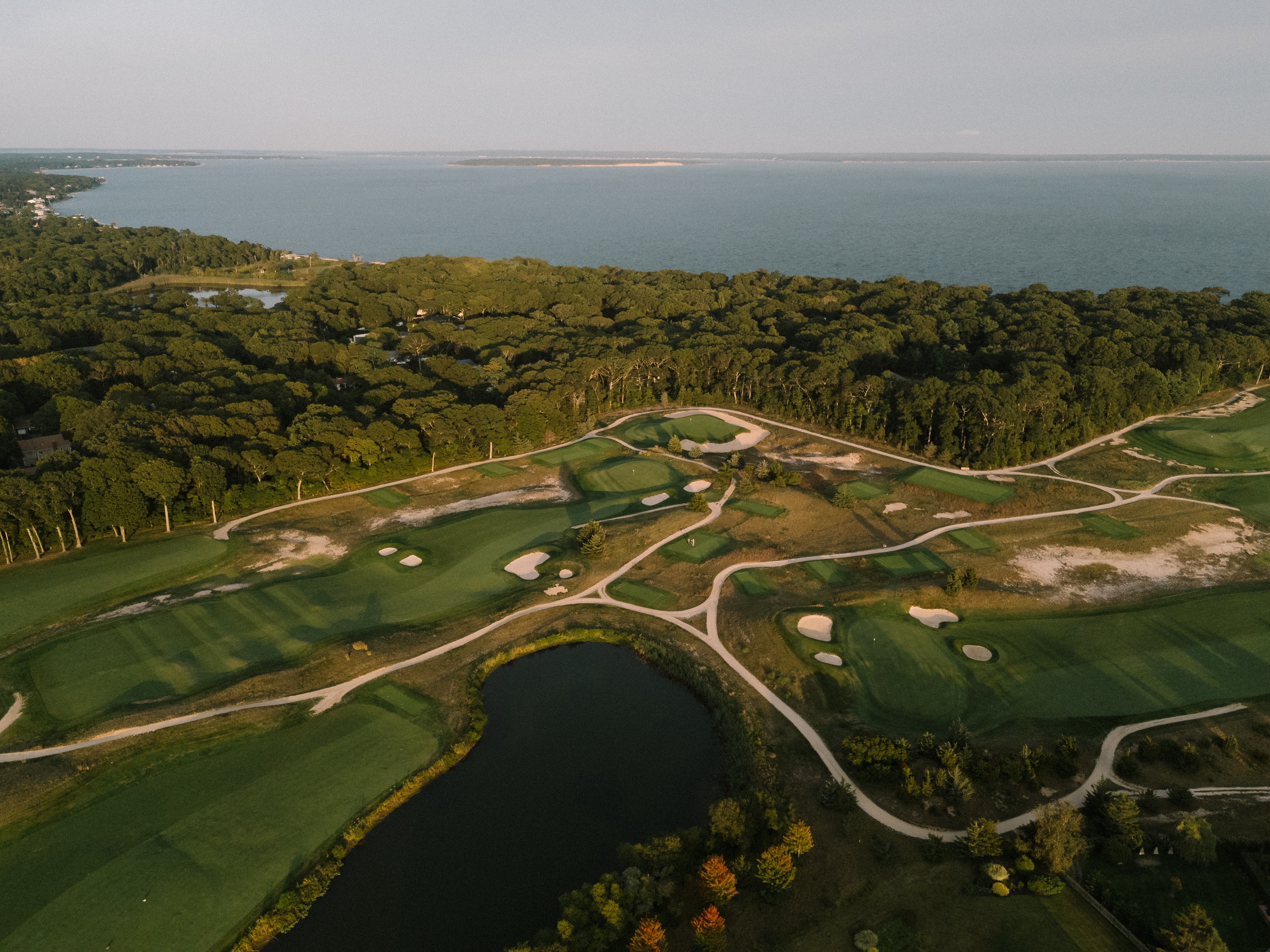 Aerial view of Laurel Links Country Club with green fairways, sand bunkers, a pond, and a tree-lined shoreline by a large body of water.