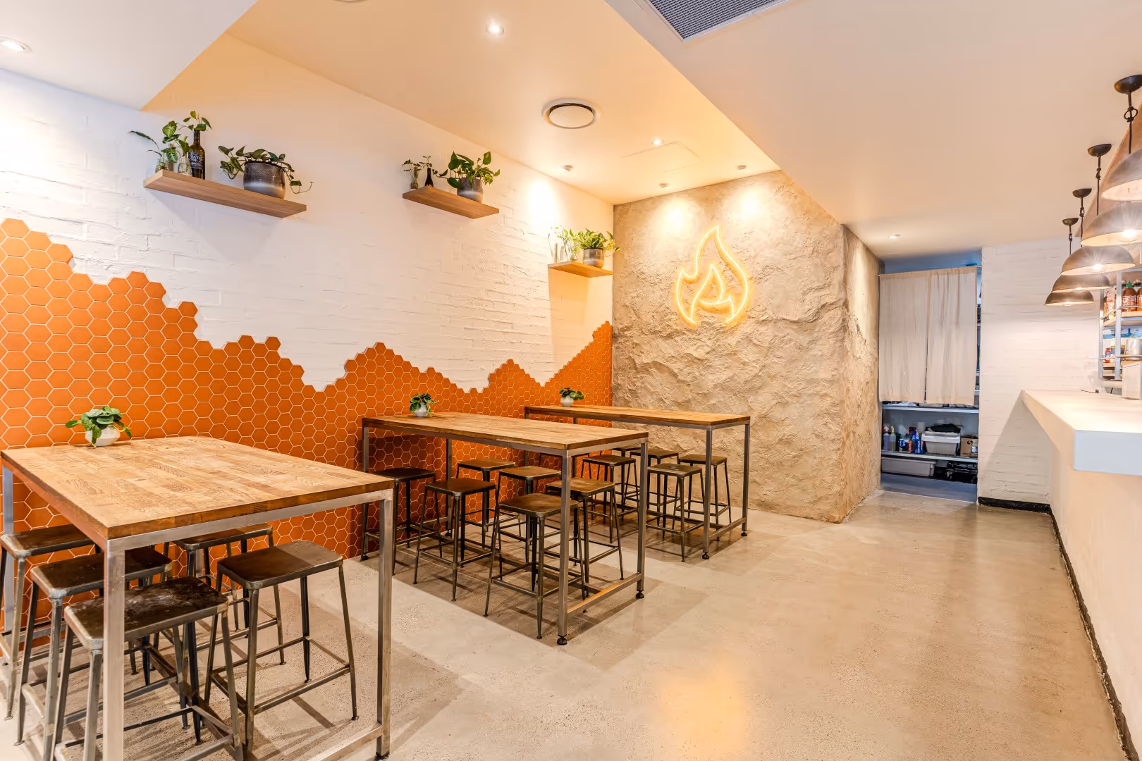 Modern café interior with wooden high tables, metal stools, orange hexagonal wall tiles, green potted plants, and a neon flame logo on a textured wall.