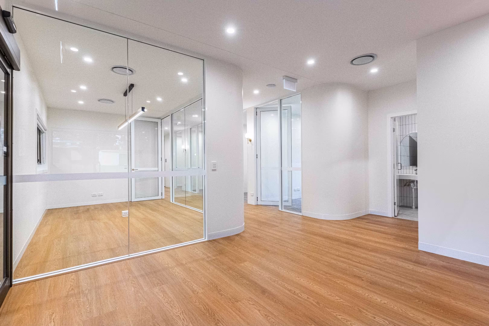 Empty modern office space with wooden floors, white walls, glass partitions, and recessed ceiling lights.
