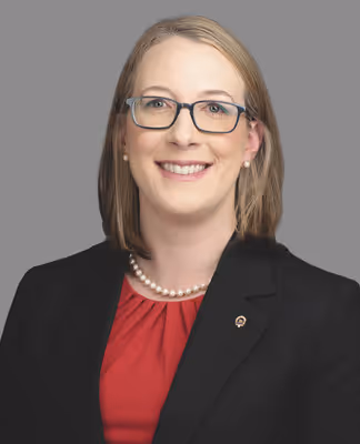 Smiling woman with glasses, pearl necklace, and pearl earrings wearing a red blouse and black blazer against gray background.