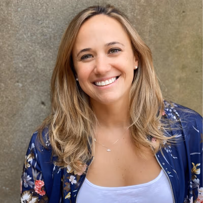Smiling woman with blonde hair wearing a floral navy jacket and white top against a textured beige background.