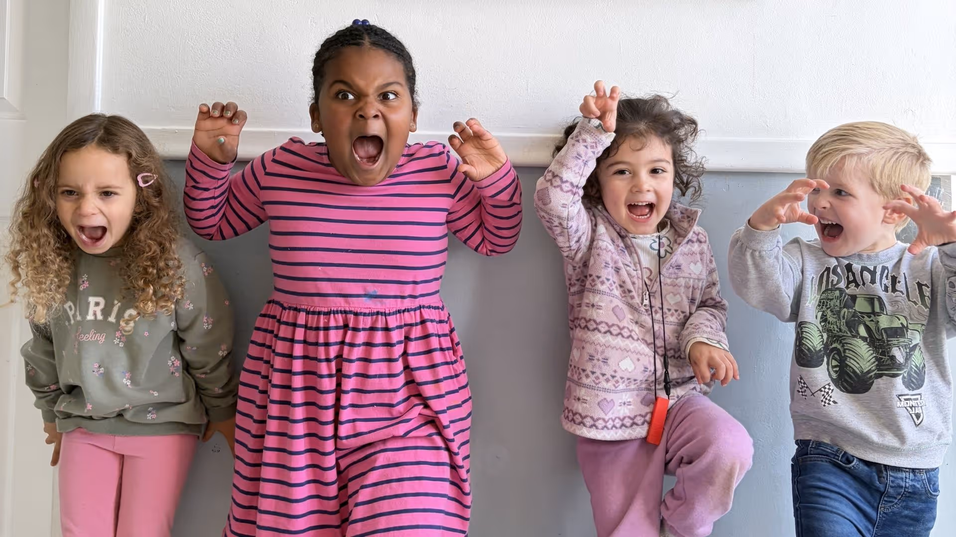 Four children standing against a wall making playful scary faces and clawing gestures.