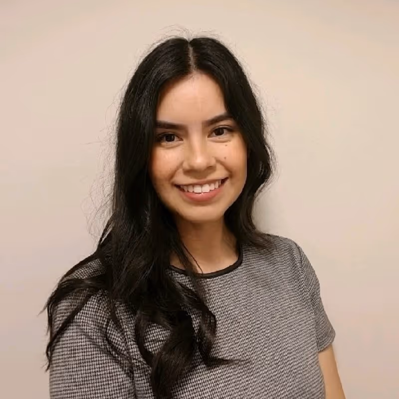 Young woman with long black hair smiling against a plain beige wall wearing a gray checkered top.