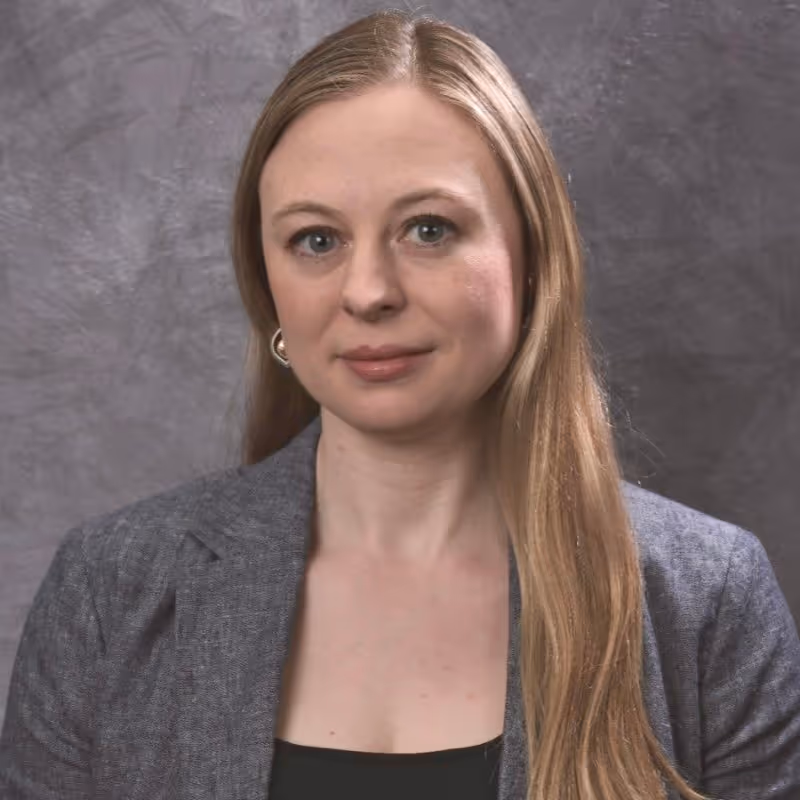 Portrait of a woman with long blonde hair wearing a gray blazer and black top against a gray textured background.