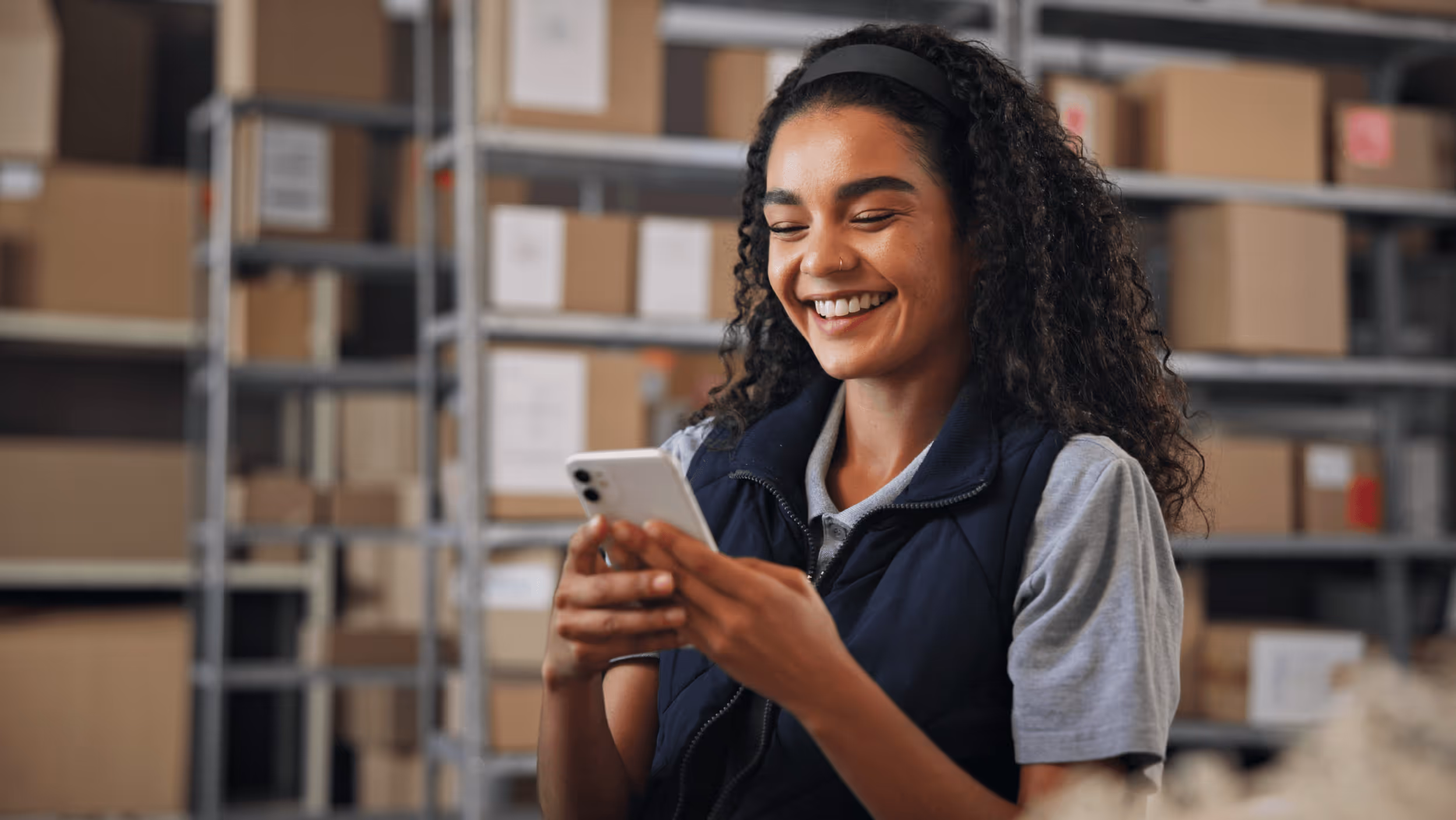 Smiling woman wearing a navy vest and gray shirt using a smartphone in a storage room with shelves of brown boxes.